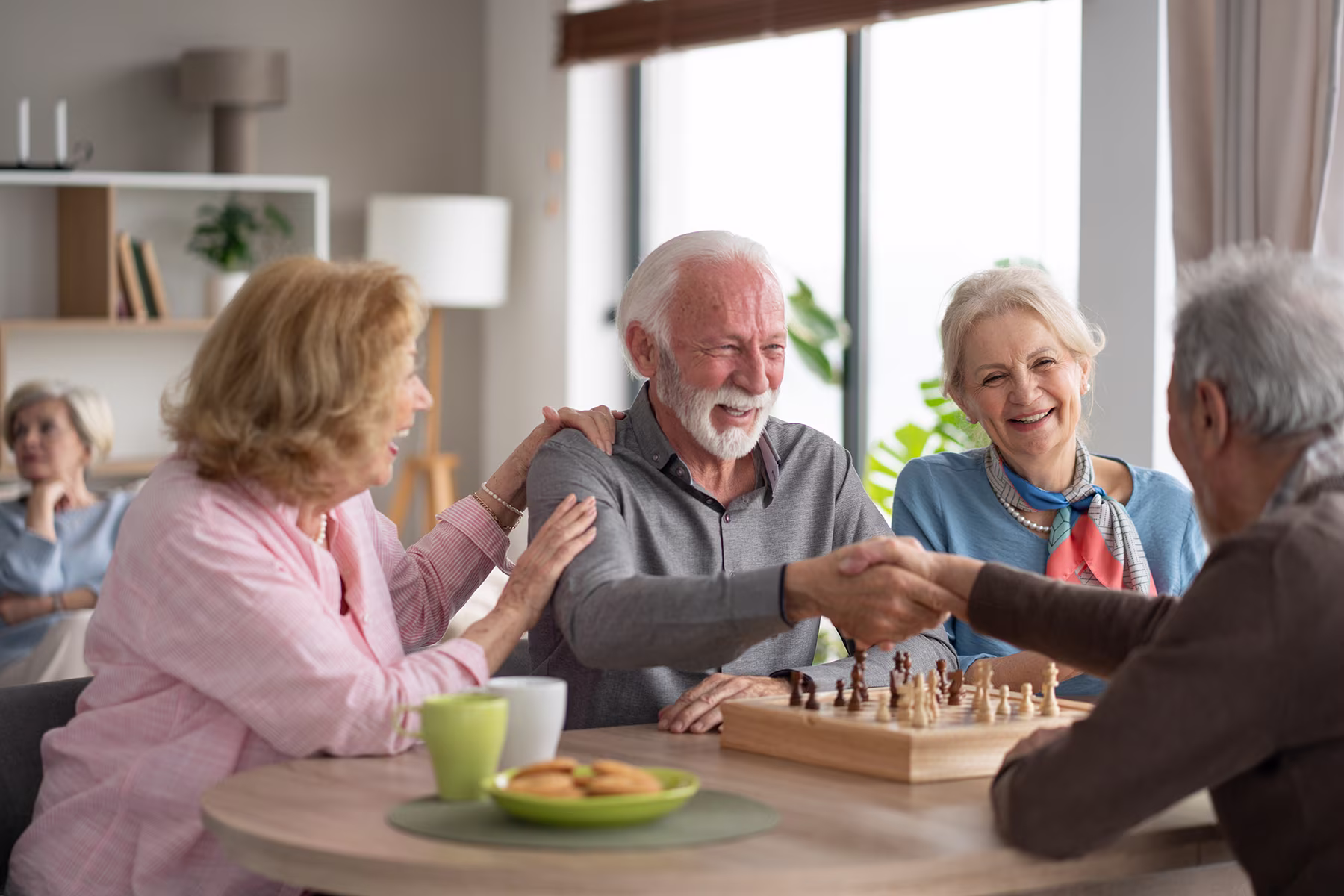 Four elderly people smiling and shaking hands over a chessboard at a round table indoors.