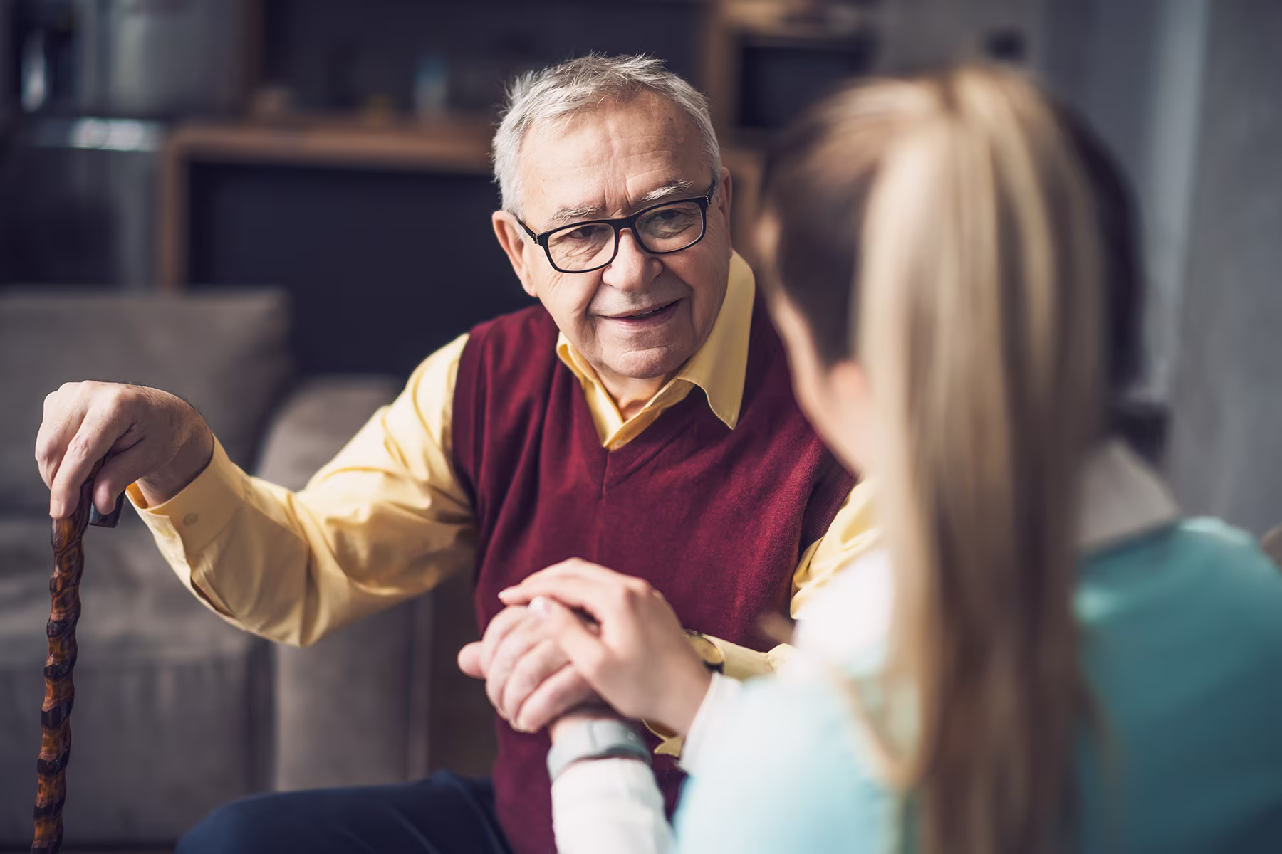 Smiling elderly man holding a cane and hands of a younger woman in a cozy room.