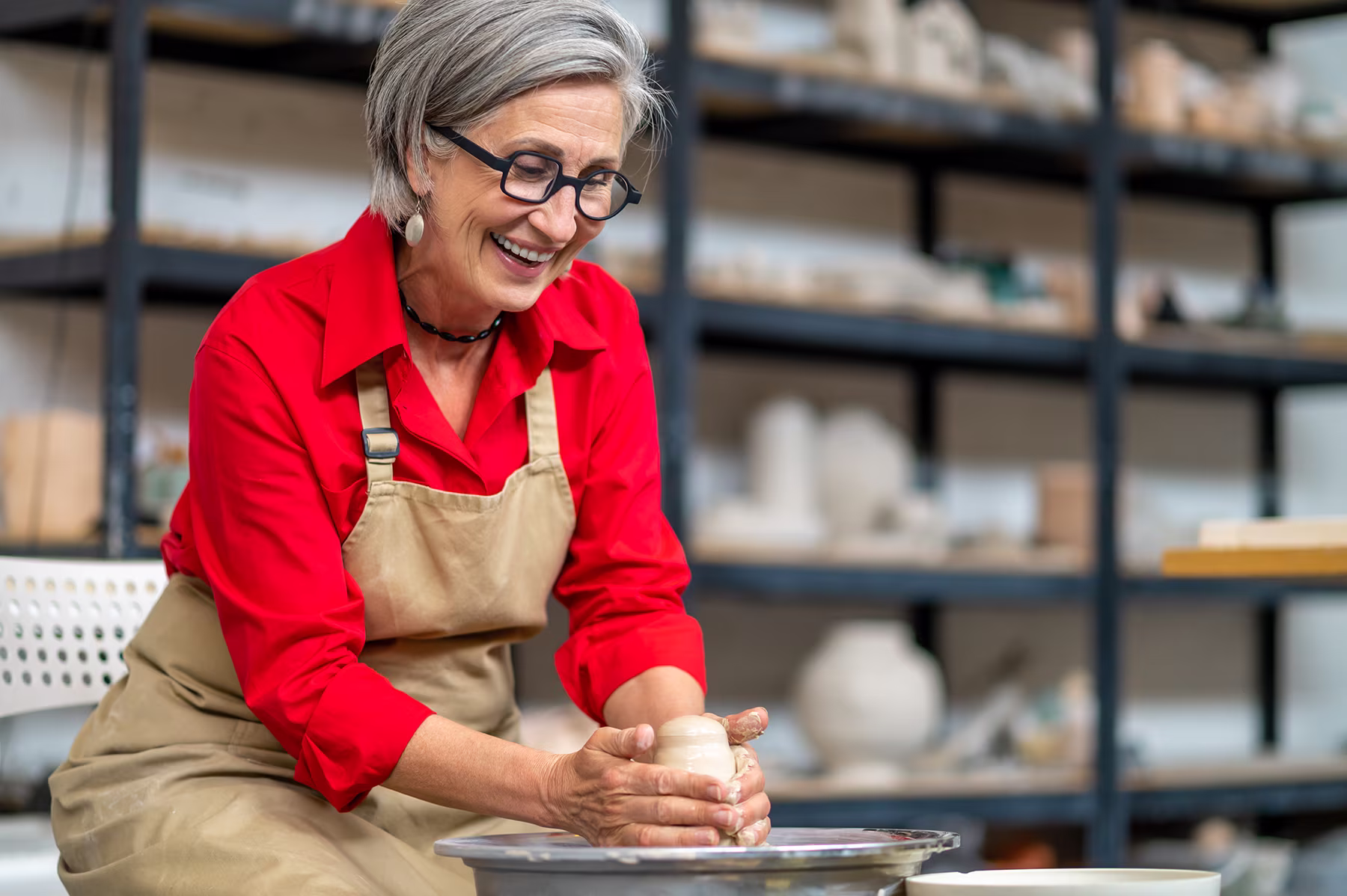 Smiling woman shaping clay on pottery wheel in a workshop with shelves of pottery behind her.