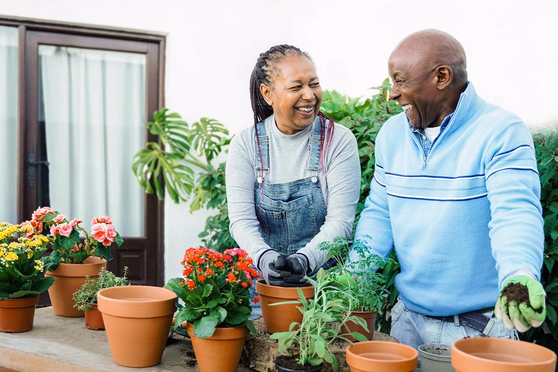 Two people smiling and gardening with potted colorful flowers and herbs on a table outside.