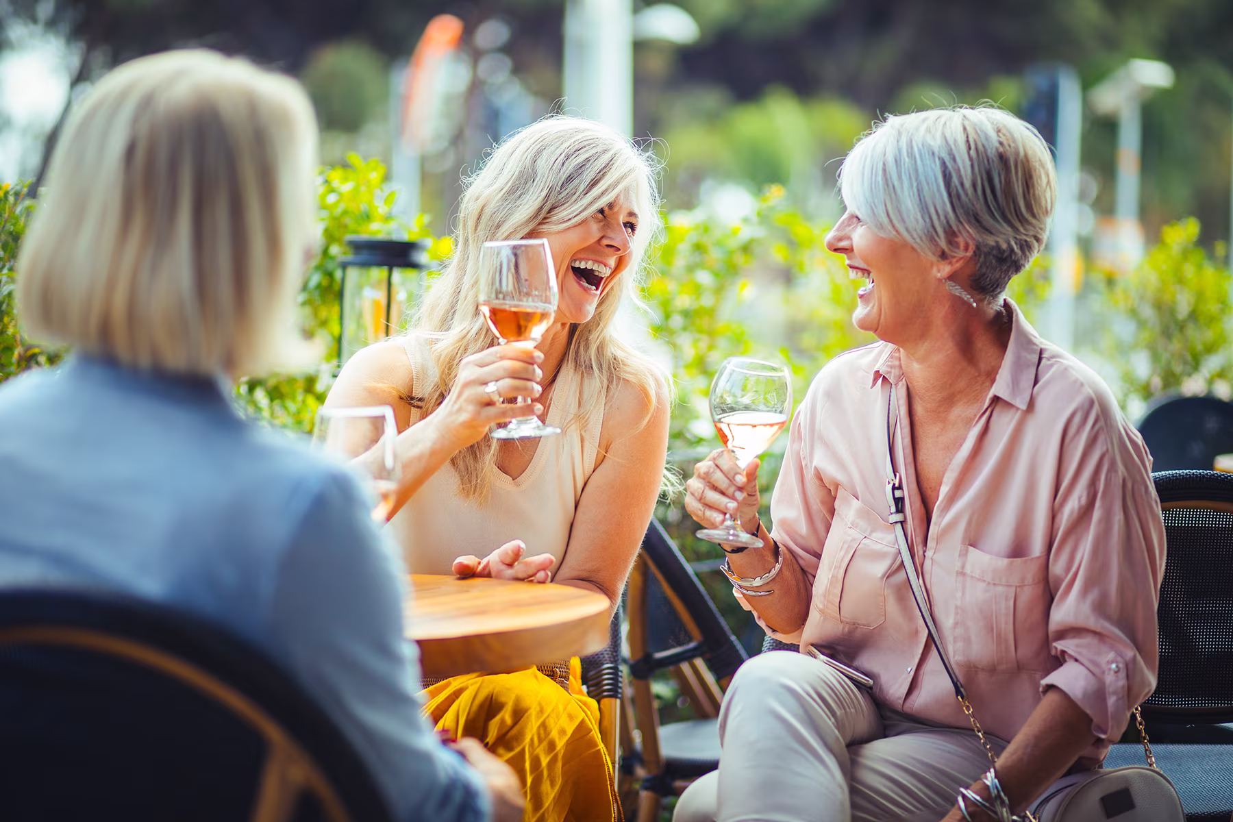 Three women outdoors smiling and toasting with glasses of rosé wine at a wooden table.