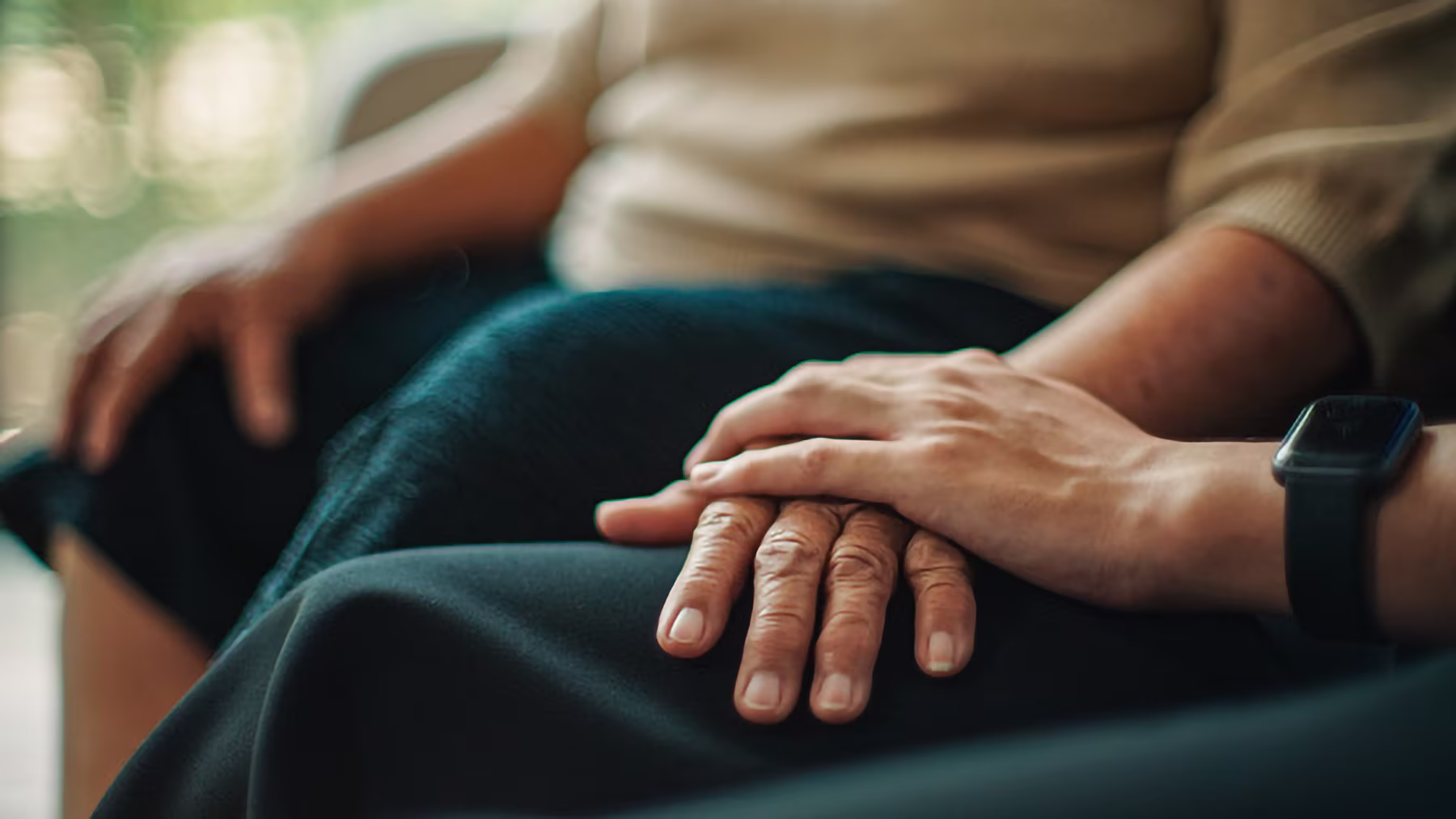 Close-up of a younger hand comforting an older hand resting on a lap.