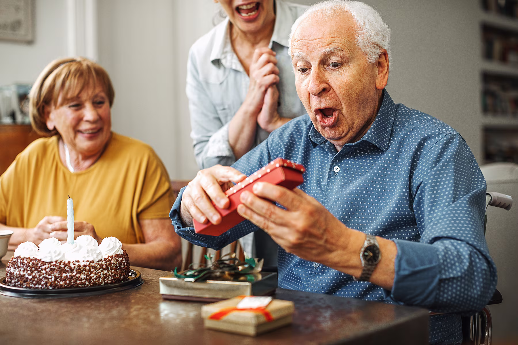 Elderly man opening a red gift box with surprised expression, two women smiling nearby with birthday cake.