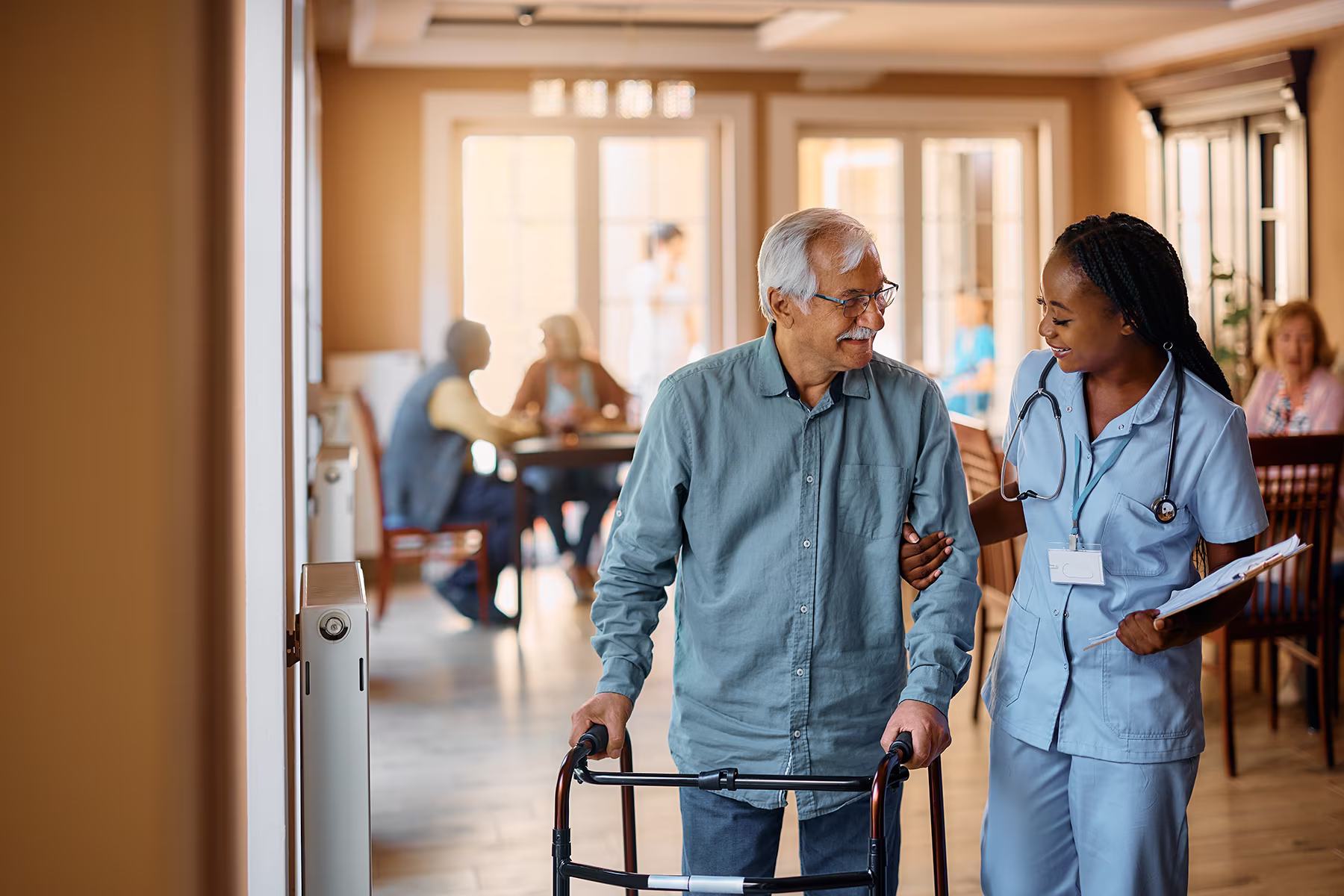 Nurse smiling while helping elderly man with walker in a care facility hallway.