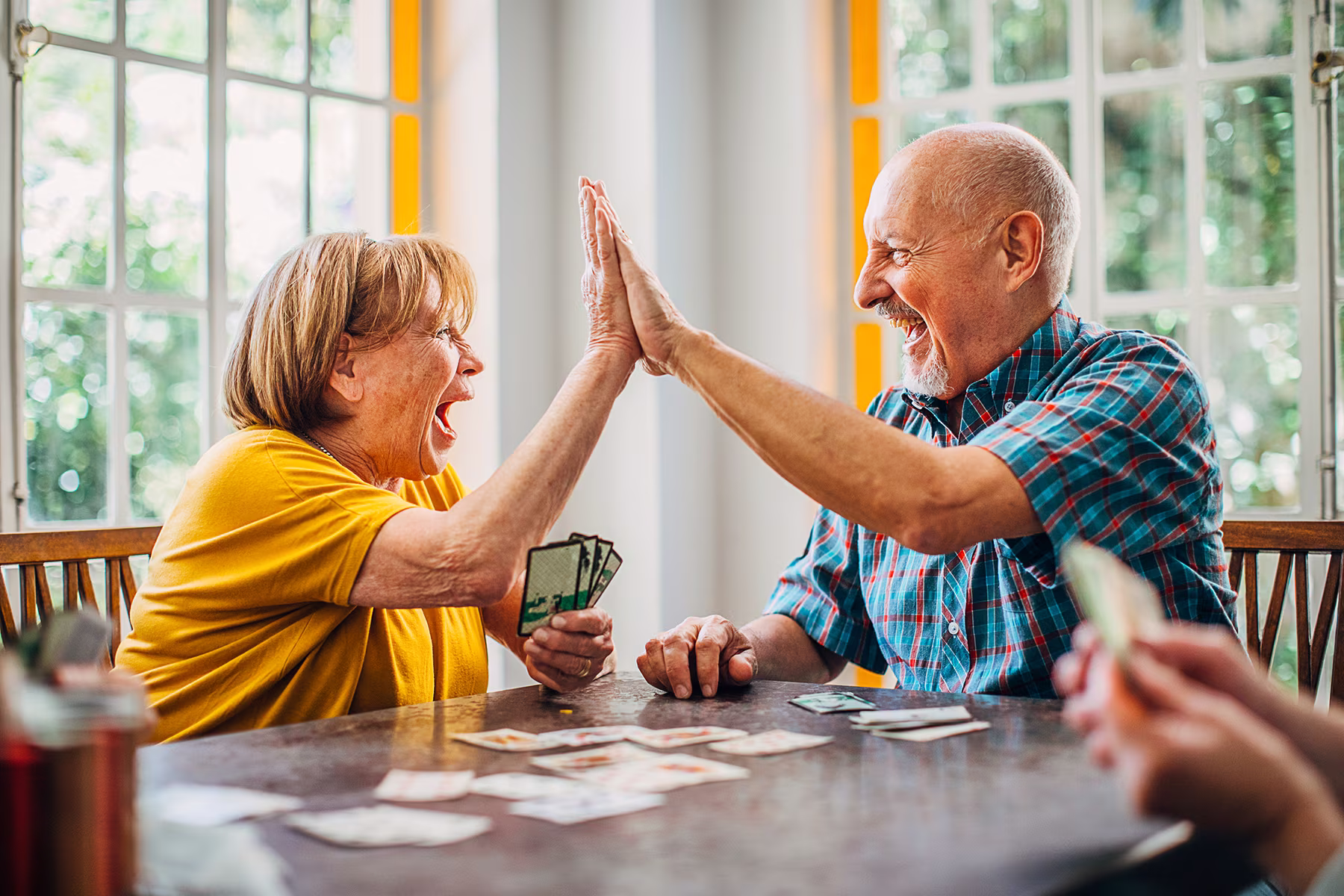 Elderly woman and man smiling and high-fiving while playing cards at a table indoors.