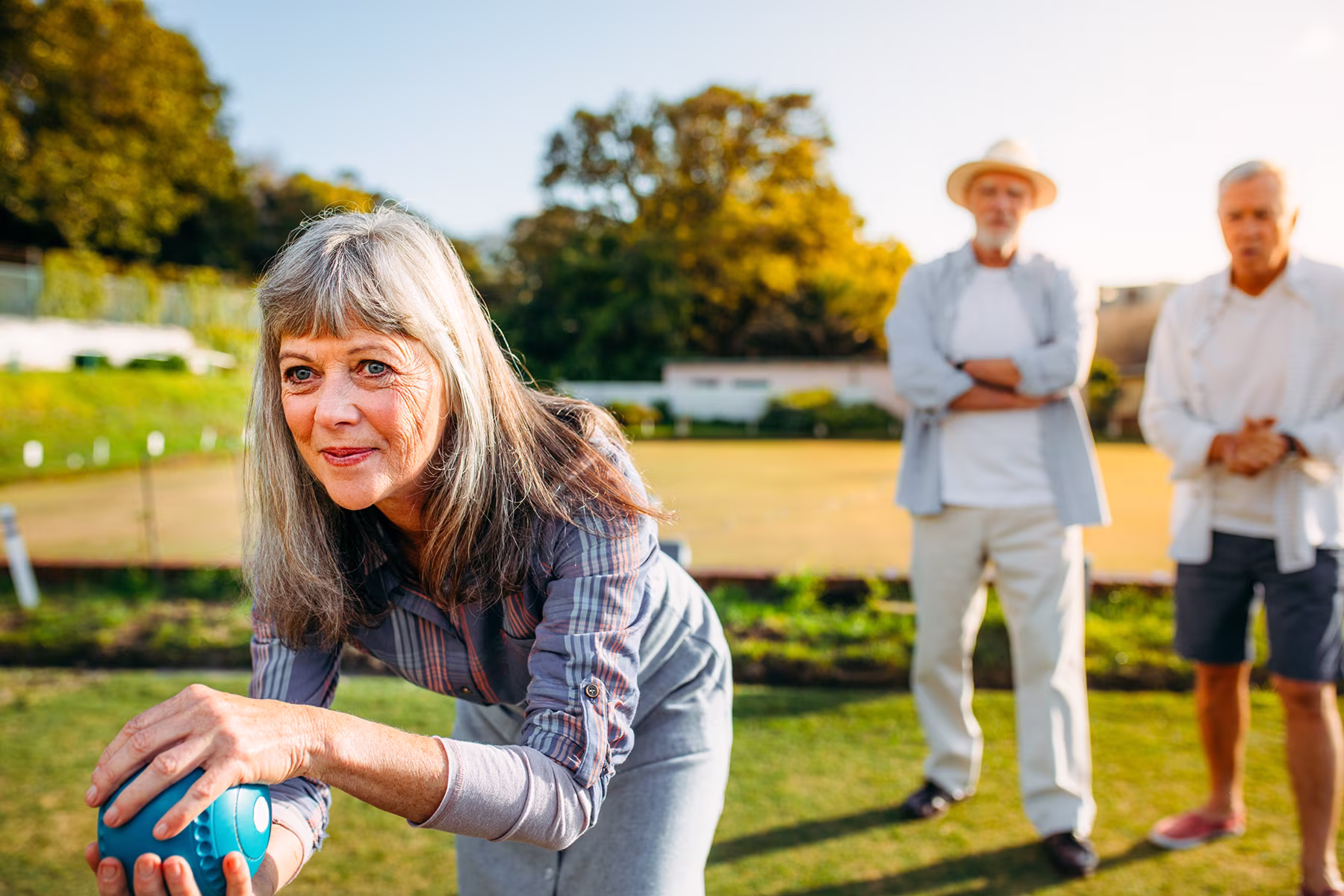 Senior woman playing lawn bowls outdoors while two older men watch in the background.