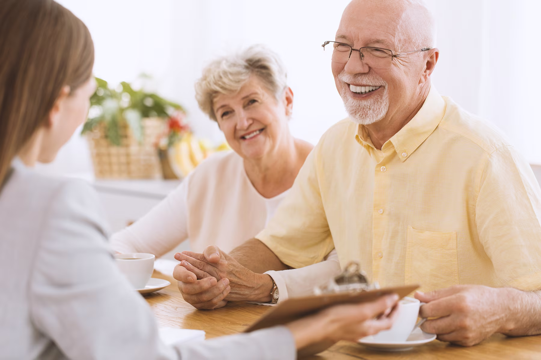 Smiling elderly couple sitting at a table having a conversation with a woman holding a clipboard.