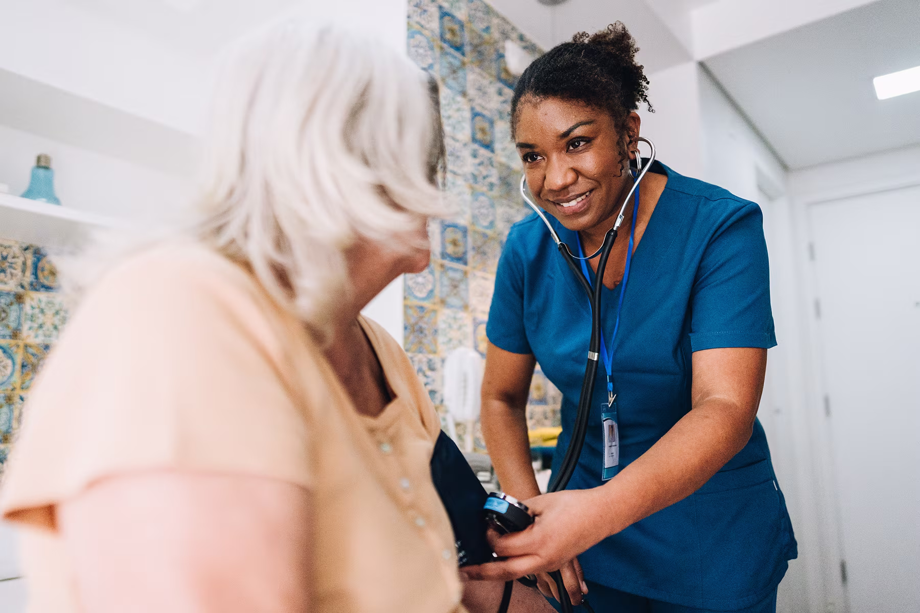 Smiling nurse in blue scrubs checking blood pressure of seated elderly woman.