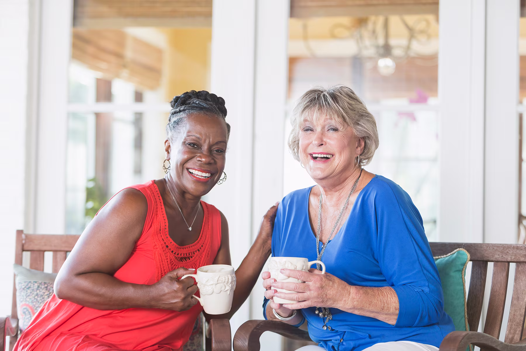 Two elderly women sitting on a bench laughing and holding mugs in a bright, cozy room.
