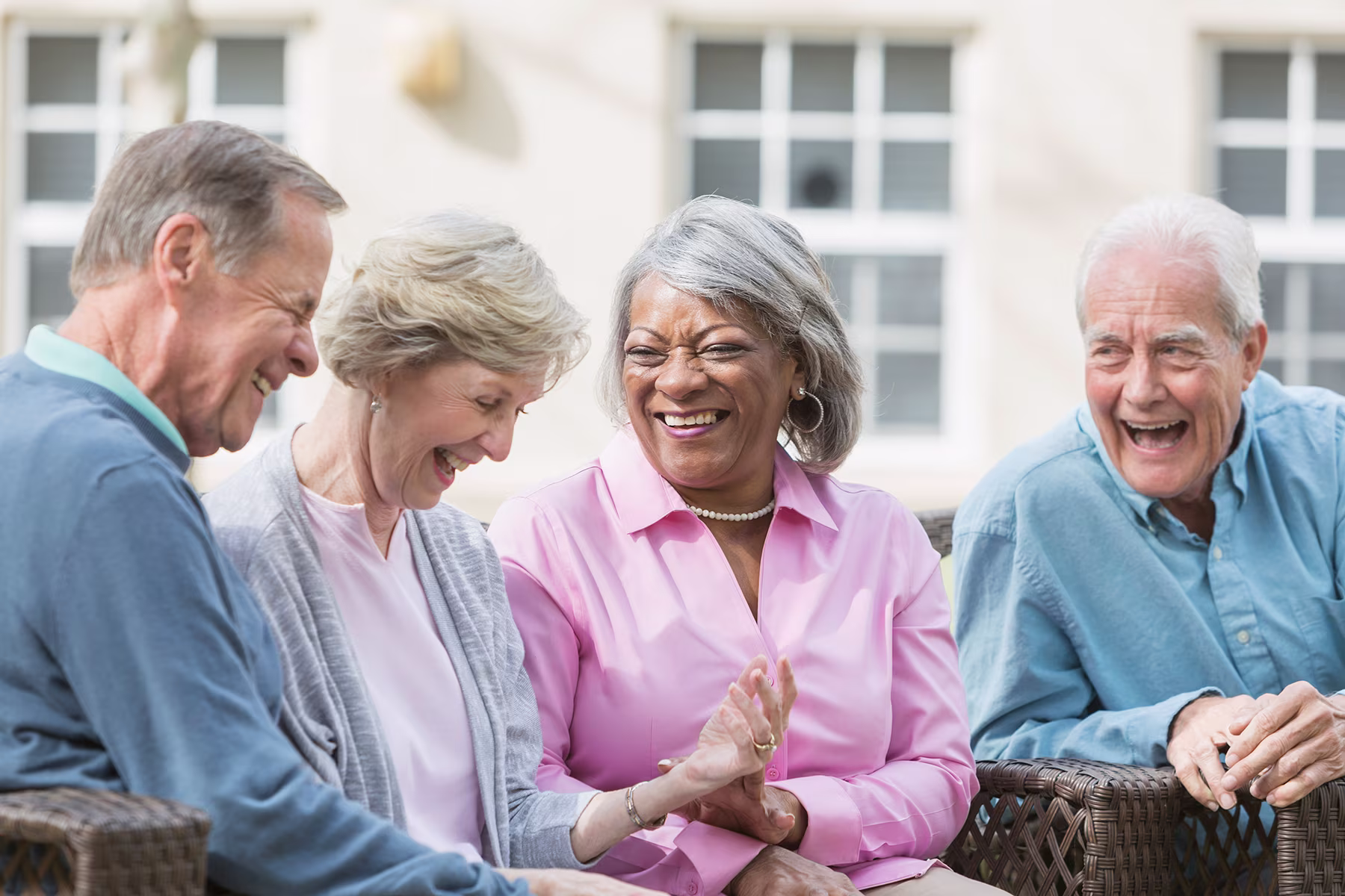 Group of four elderly friends sitting outdoors, smiling and laughing together on wicker chairs.