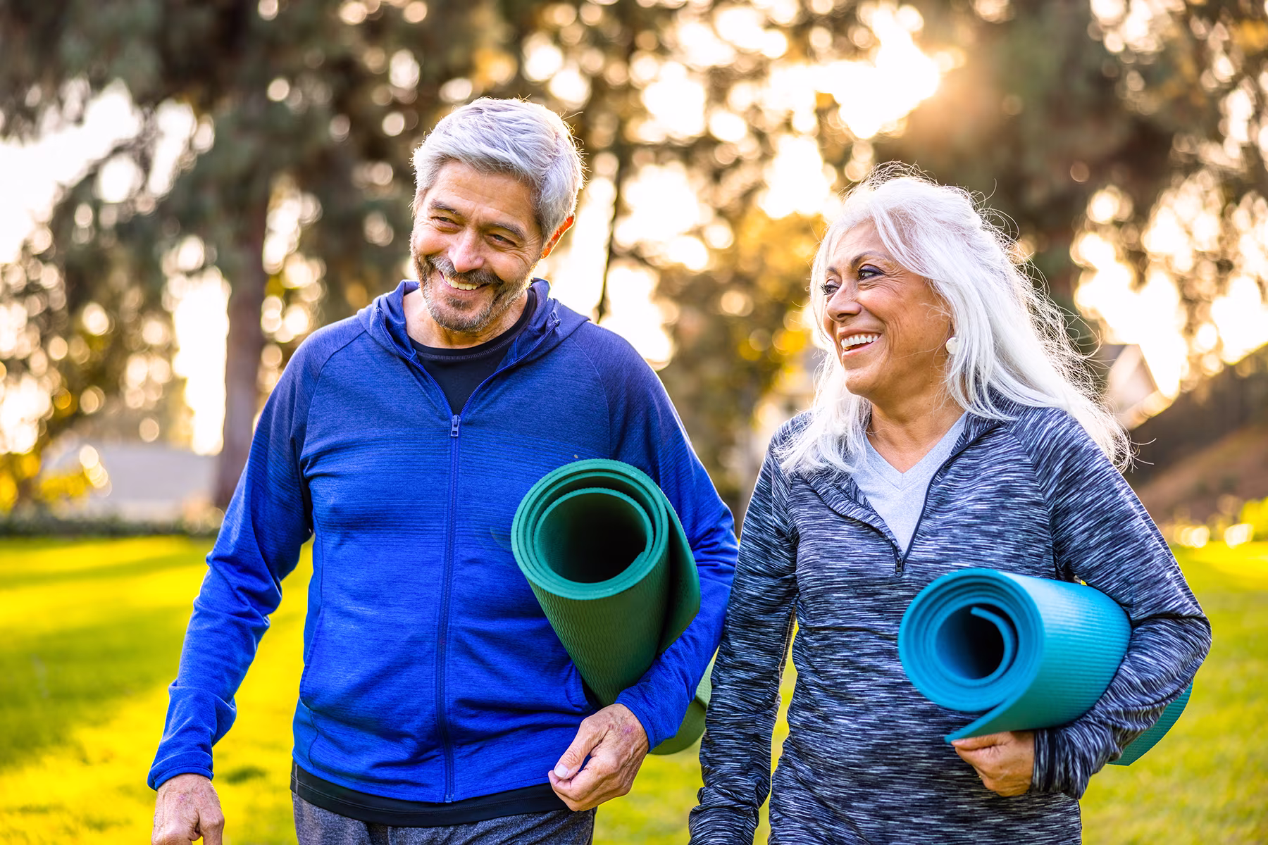 Smiling senior couple walking outdoors, each carrying a rolled yoga mat under their arm.