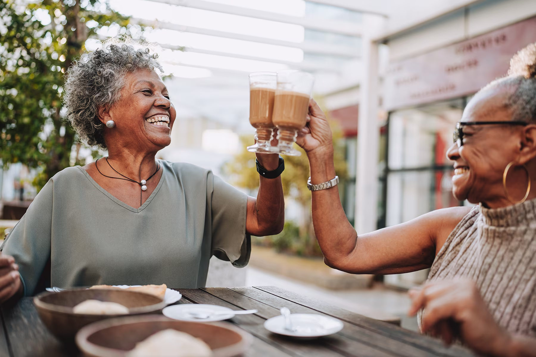 Two elderly women smiling and clinking glasses of coffee while sitting at a wooden table outdoors.