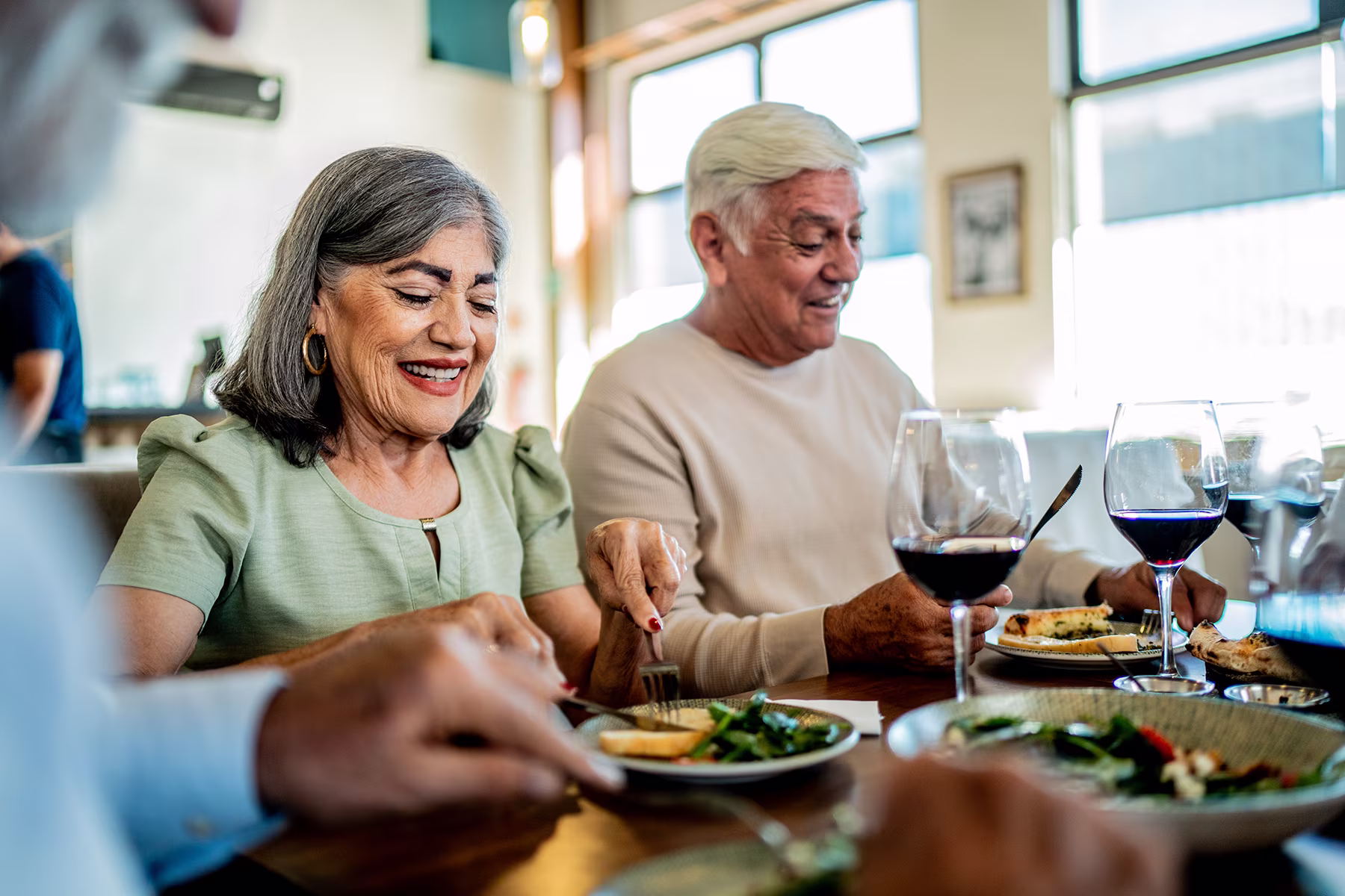 Smiling elderly couple enjoying a meal with wine at a bright restaurant table.