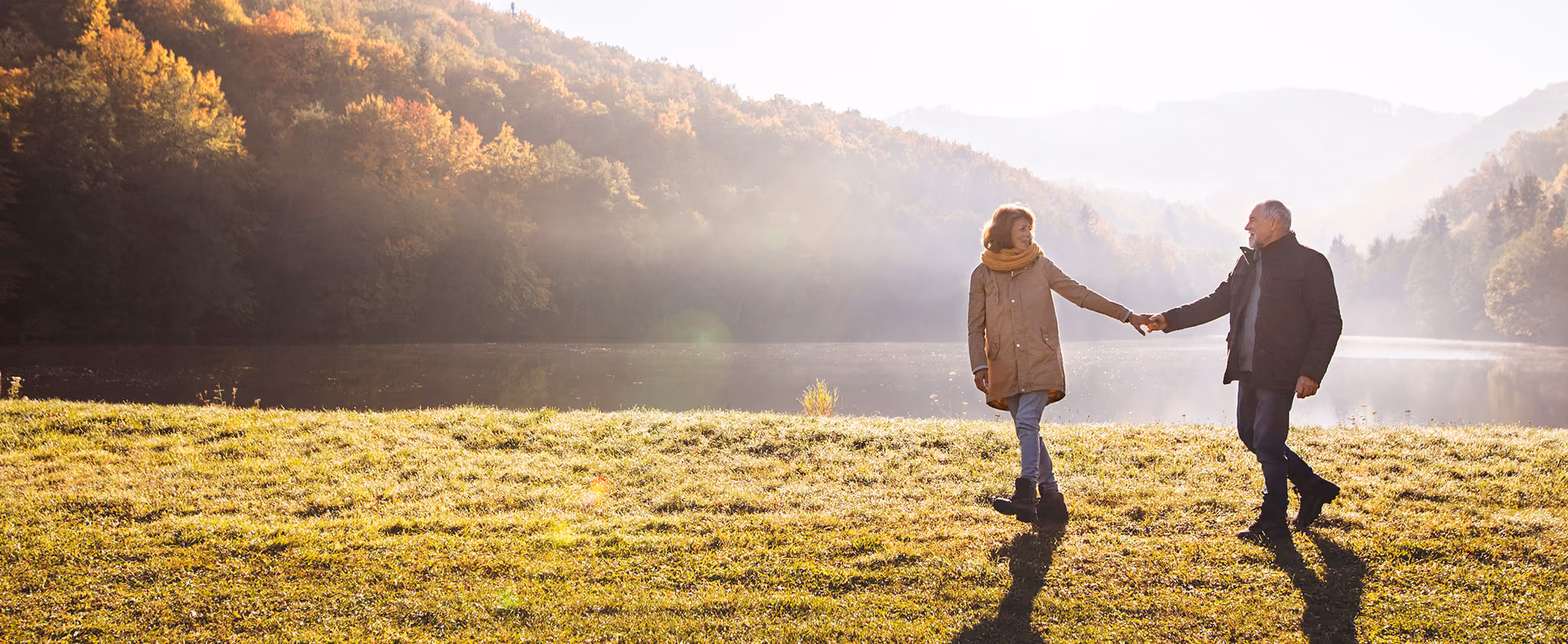 A couple holding hands and walking on grass near a calm lake surrounded by autumn trees and hills.