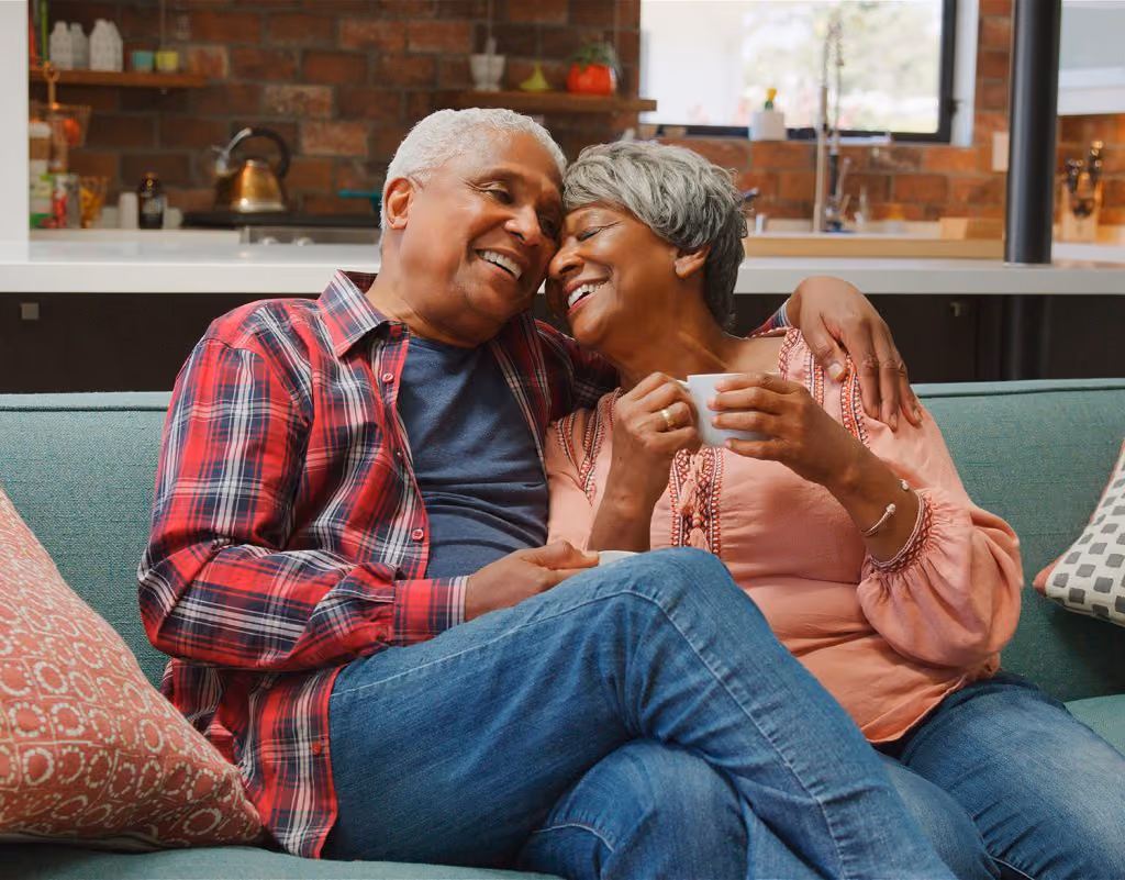 Smiling elderly couple sitting close on a couch, holding coffee cups and sharing a warm moment.