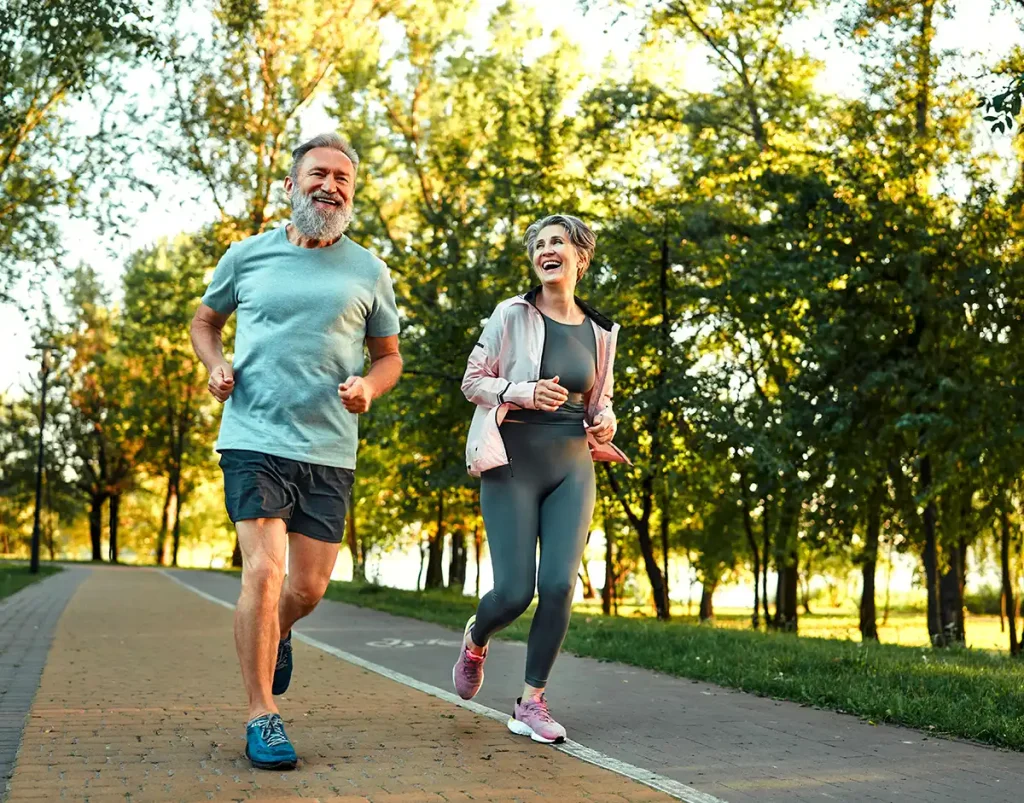 Smiling older man and woman jogging together on a tree-lined park path in daylight.