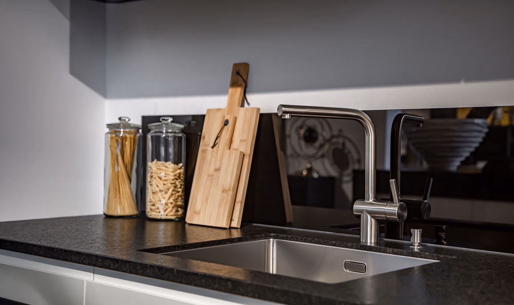 A modern kitchen sink with a faucet and cutting boards beside two jars of pasta on the counter.