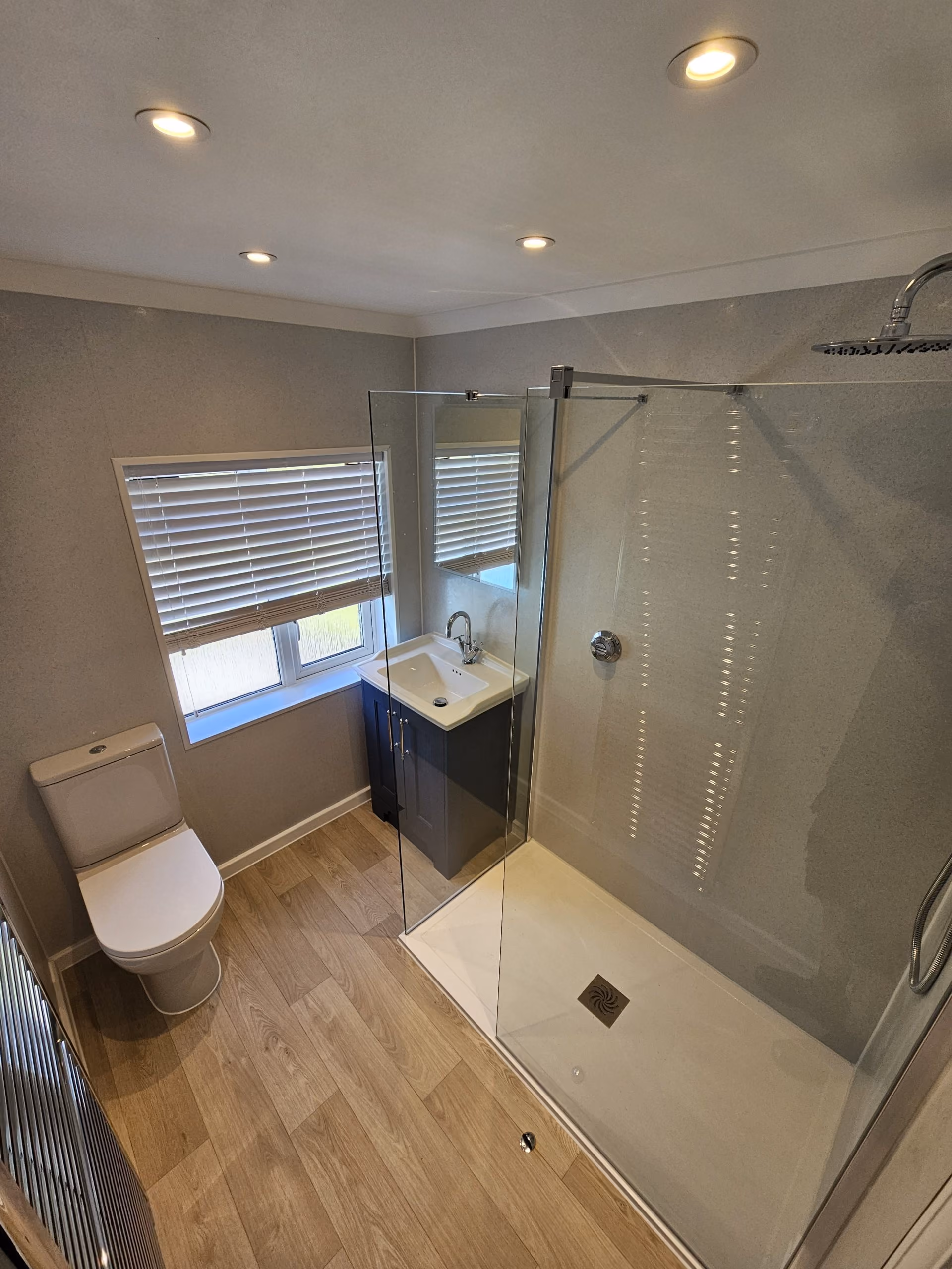 A modern bathroom with a glass shower, toilet, and sink under a window with blinds.