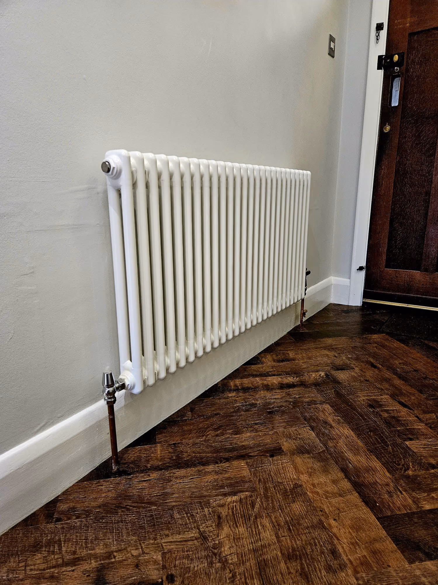 A white radiator is attached to a light-colored wall on a wooden floor.