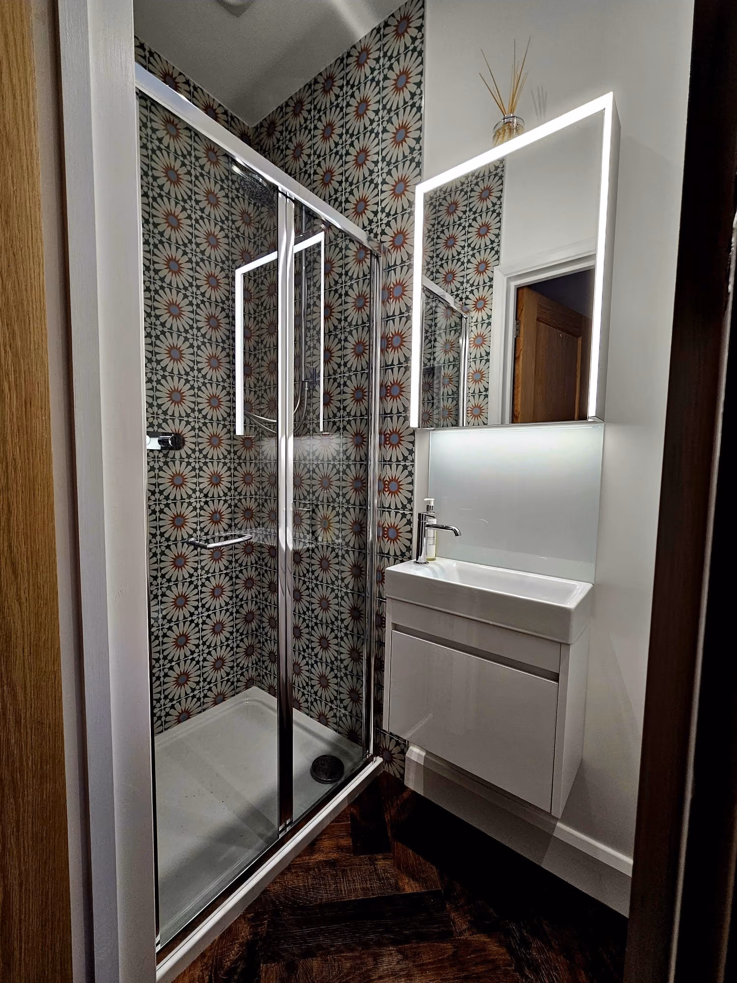 Modern bathroom with a glass shower, geometric patterned wall, illuminated mirror, and white sink.