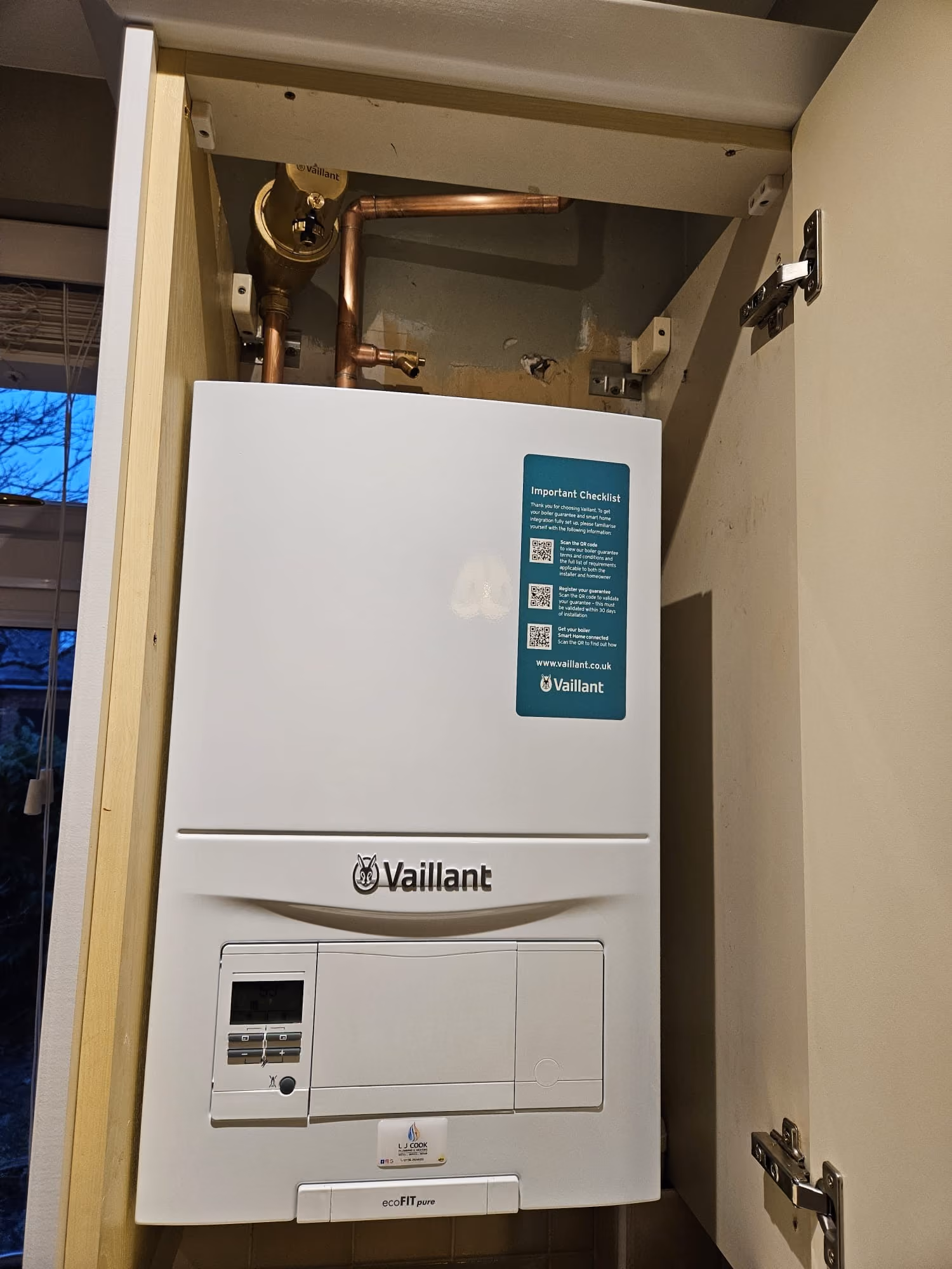 White boiler unit with visible copper pipes inside a cupboard, door open.