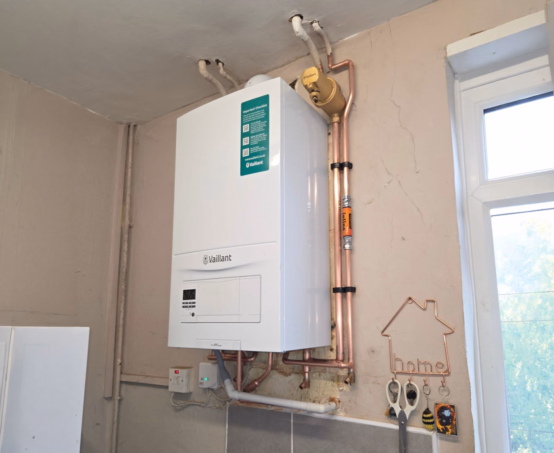 Wall-mounted boiler with copper pipes and a small house-shaped decoration nearby on the wall.