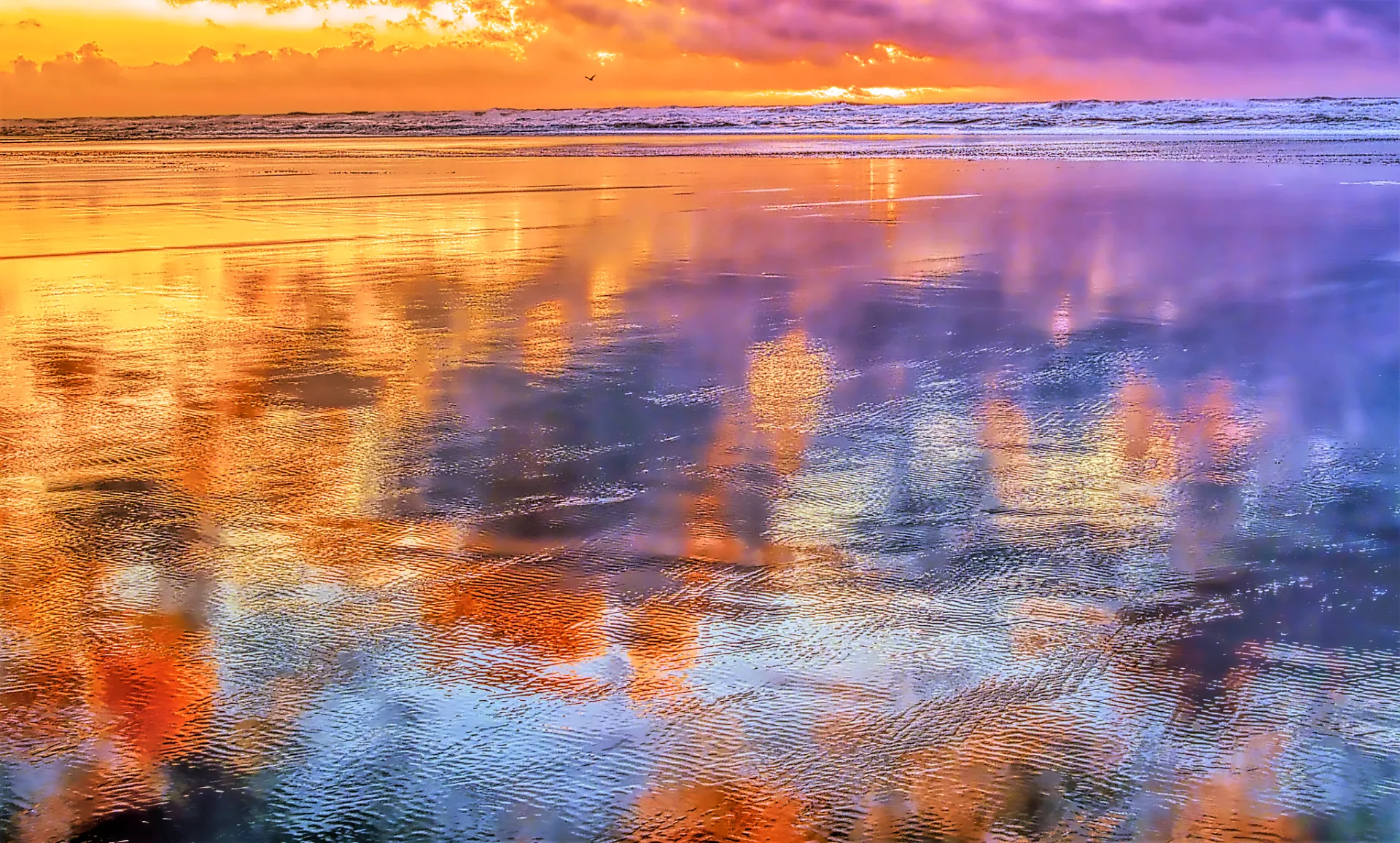 Vibrant sunset over ocean waves reflecting bright orange, purple, and blue colors on wet sand.