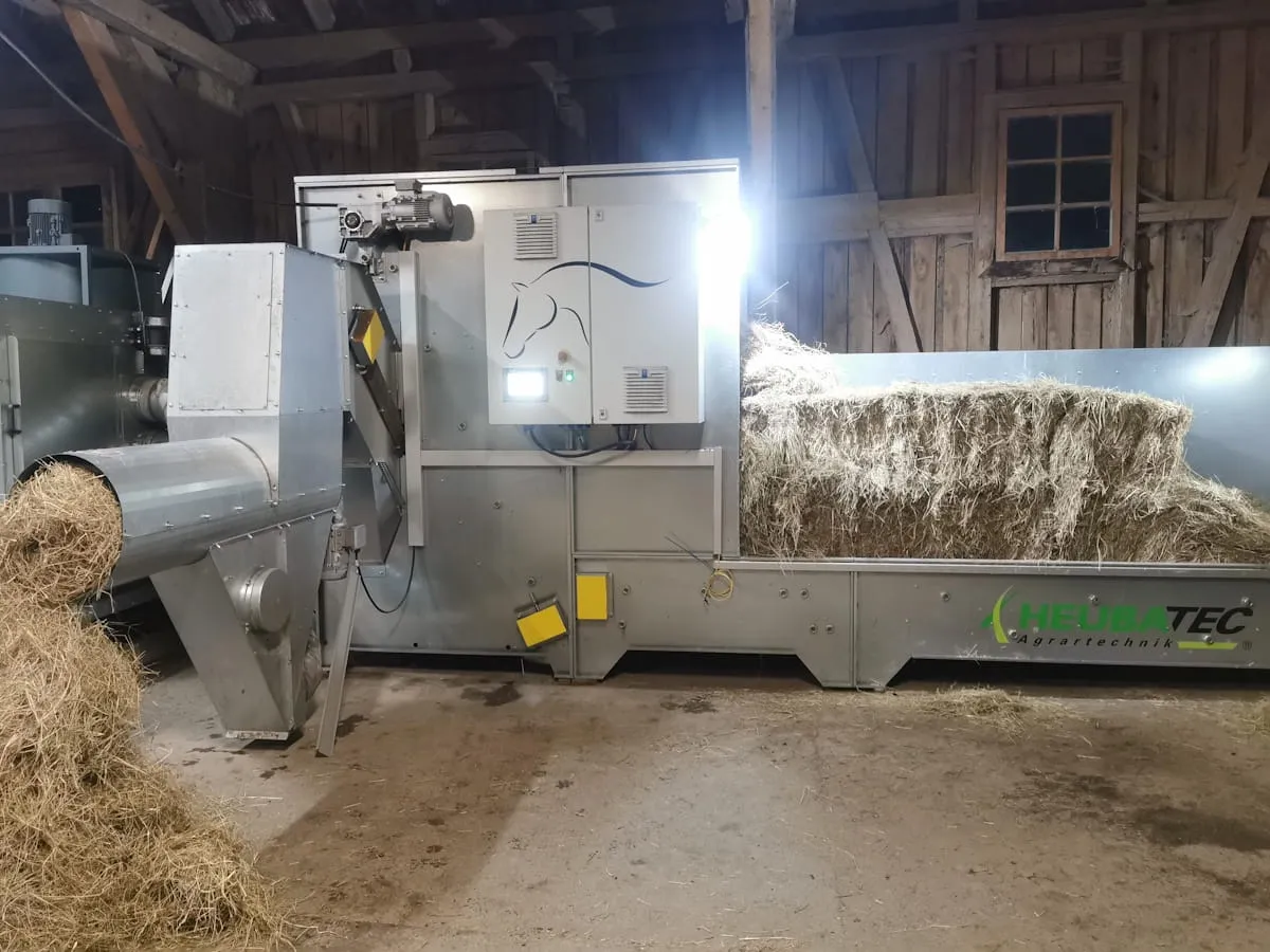 Indoor agricultural machine processing hay with bales stacked on a metal platform in barn setting.
