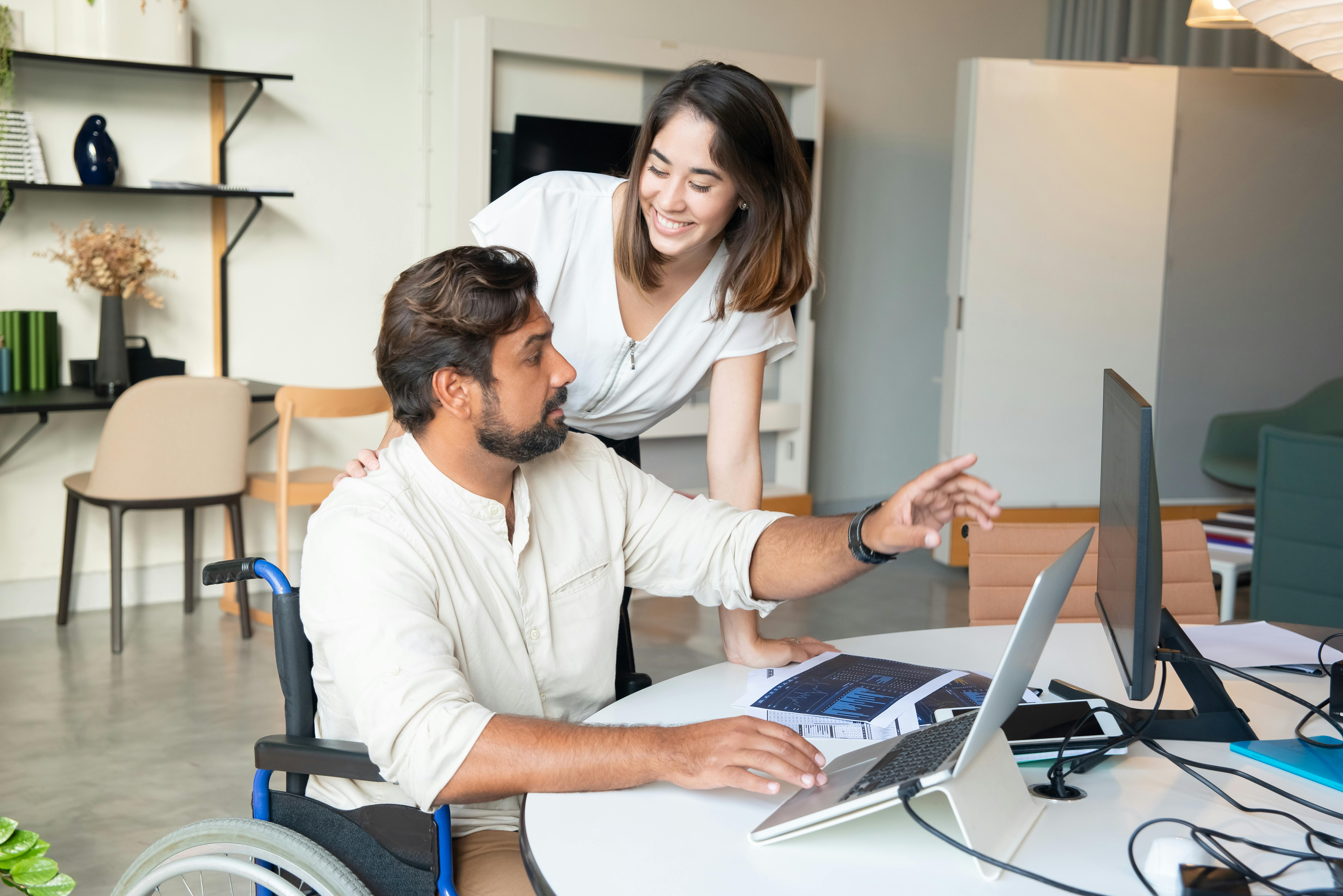 A female broker assists a colleague sitting at a desk looking at a computer