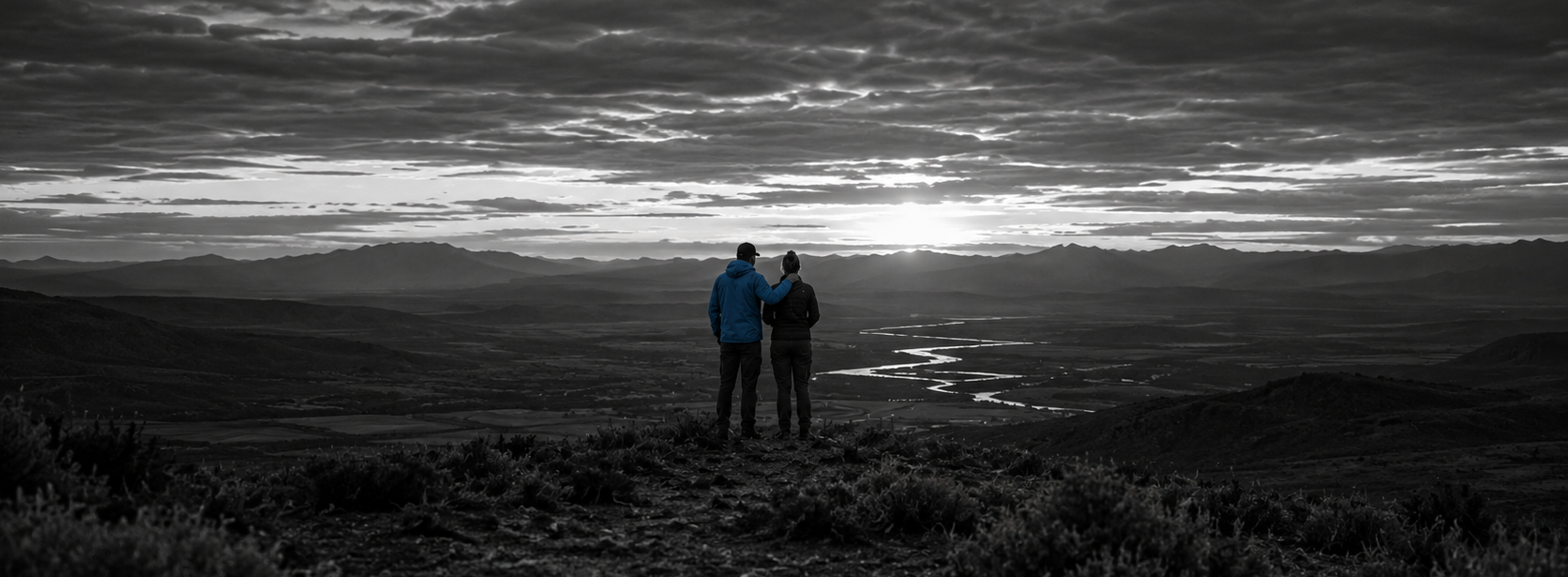 Two people stand on a mountain overlook at sunset, one in a blue jacket with an arm around the other, looking out over a river valley.