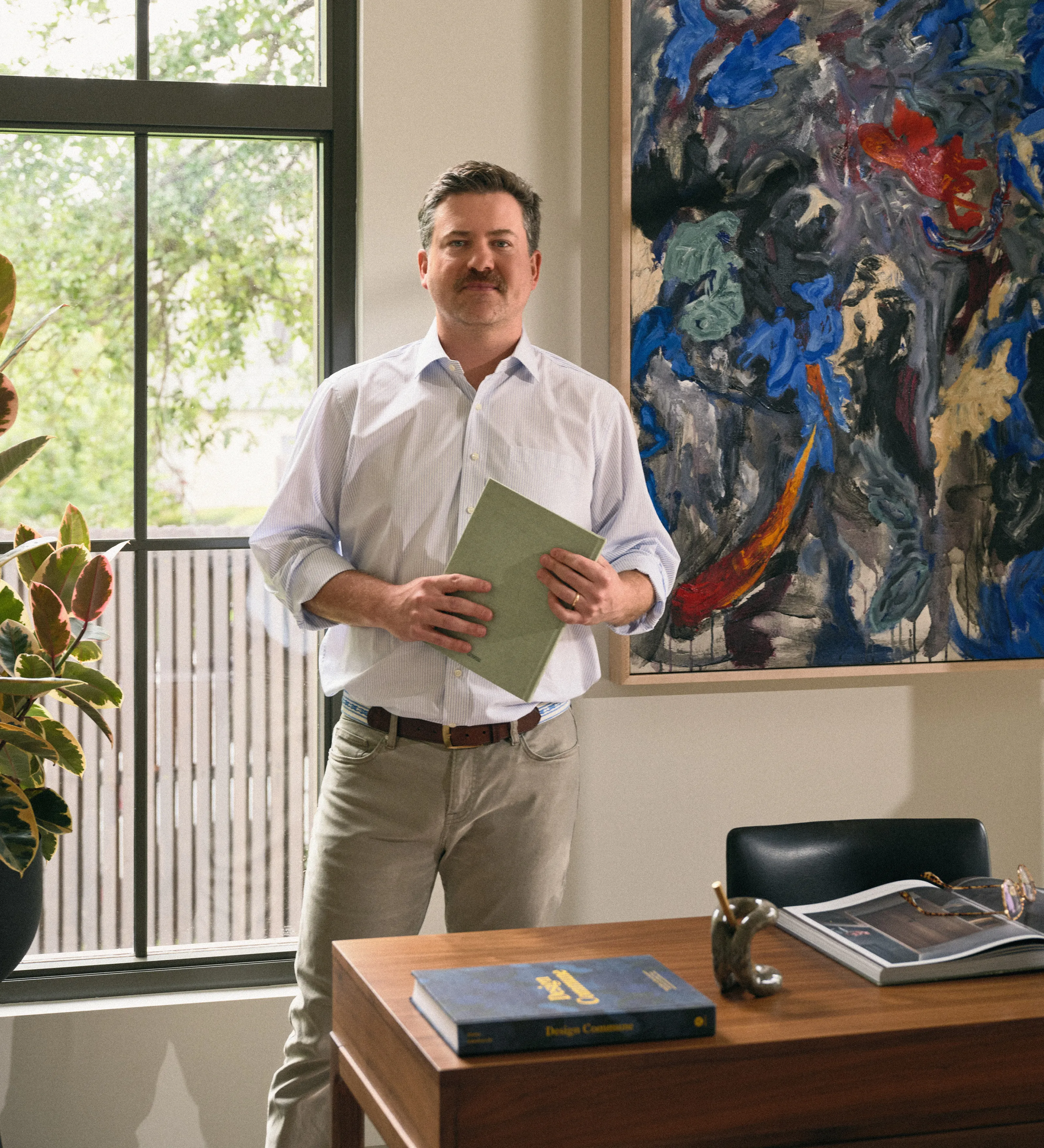 Man with mustache in casual clothes holding a folder standing by a desk with books and glasses.