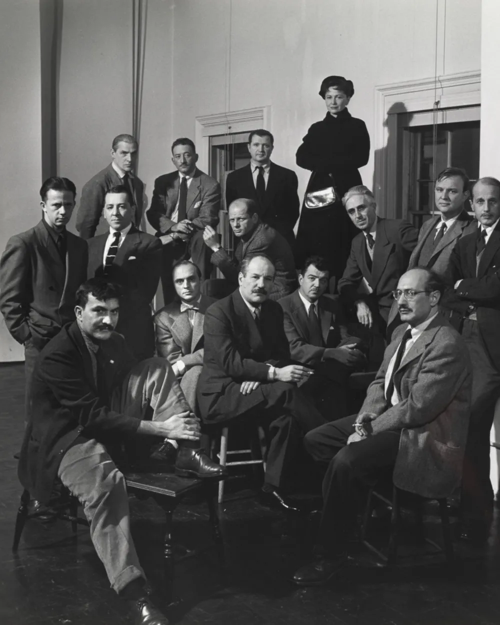 Group of sixteen people, mostly men, in formal attire posing in a room with a plain wall and windows.