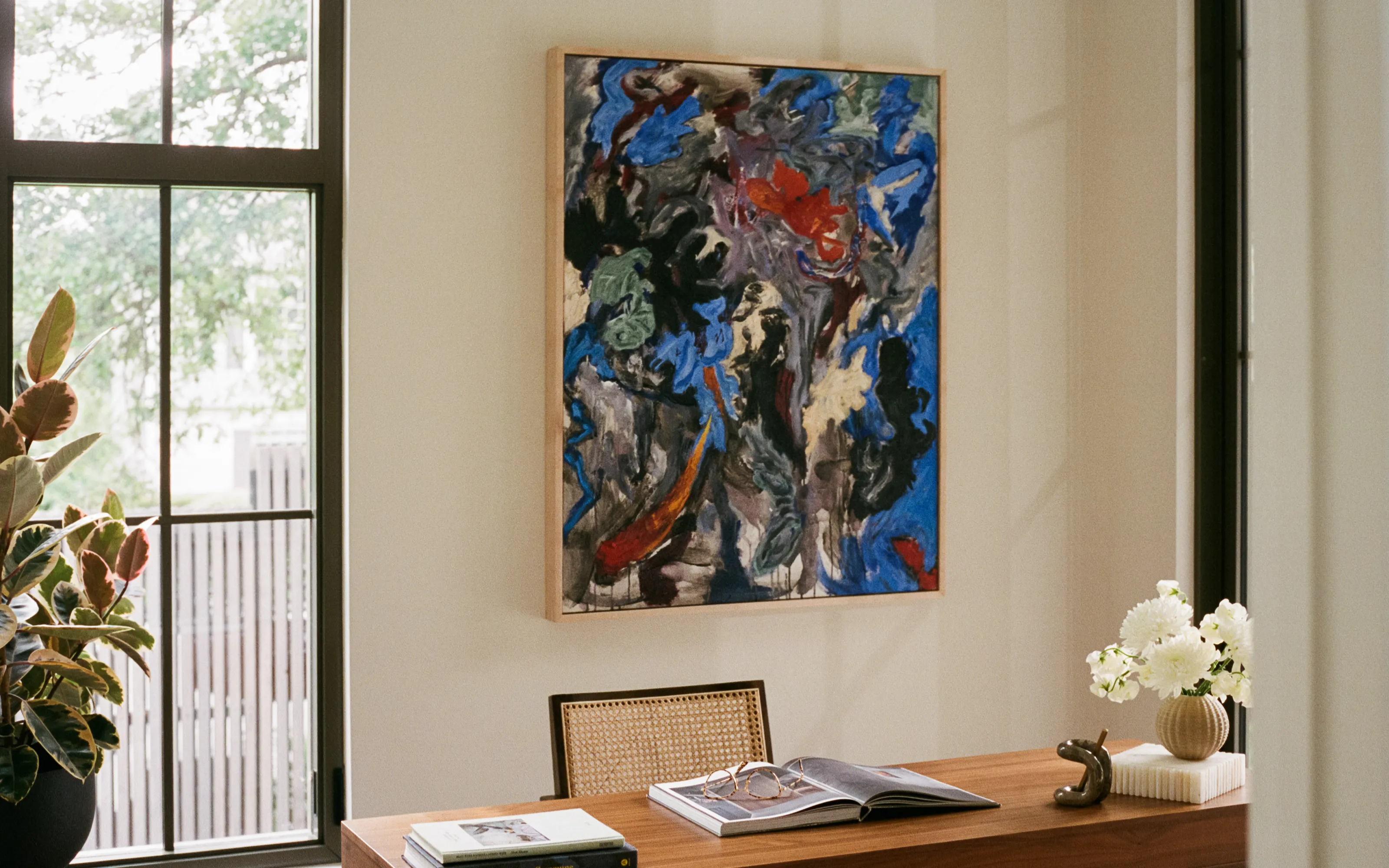 Cozy living room with beige sofa, cushions, wooden coffee table, plants, abstract blue painting, and wood beams.