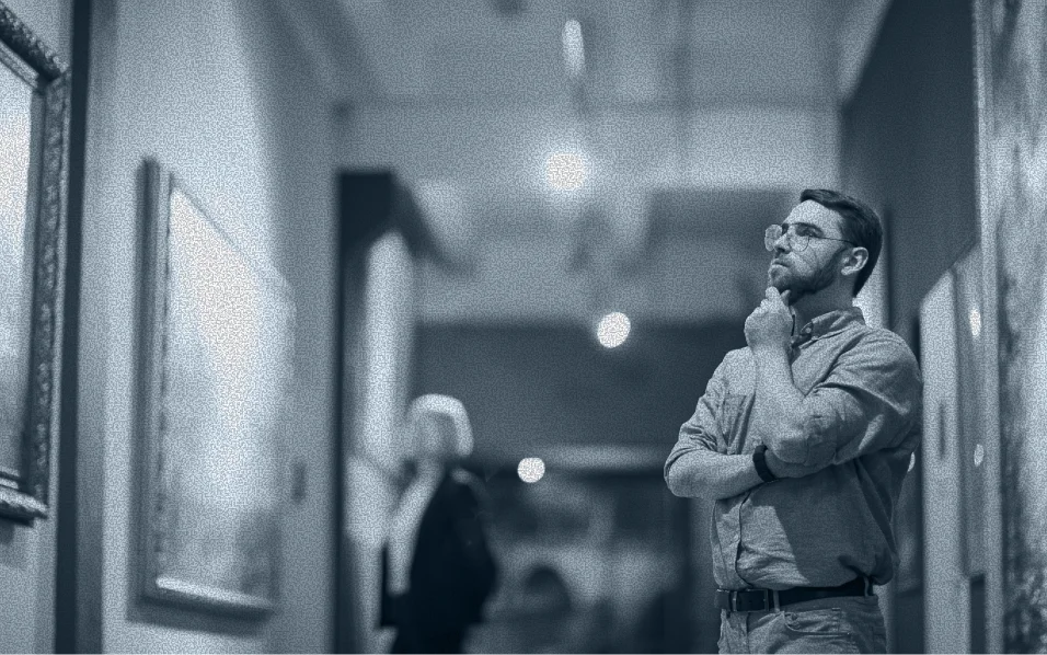 Man with glasses thoughtfully observing artwork in a gallery with blurred visitors in the background.