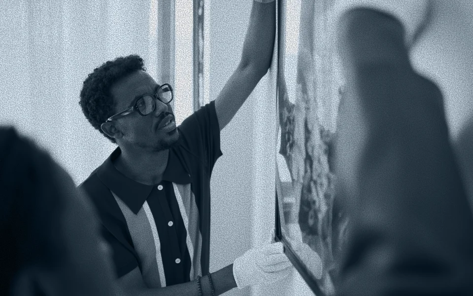 Man wearing glasses and white gloves adjusting a framed artwork on a wall.