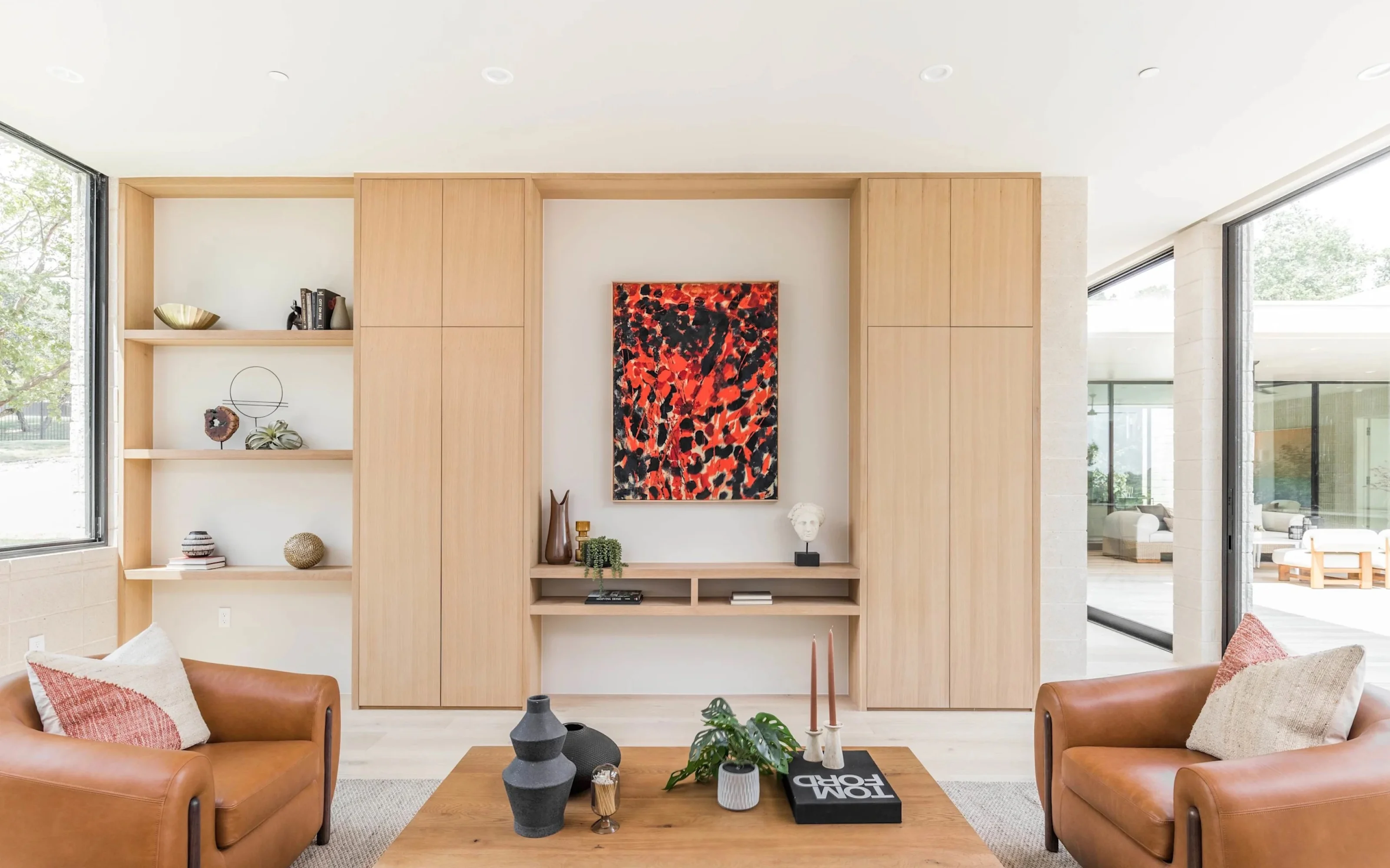 Modern living room with tan leather chairs, wooden shelves, abstract red and black painting, and large windows.