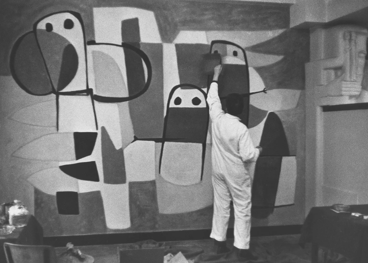 Artist in white coveralls painting a large abstract mural with geometric shapes on a wall.