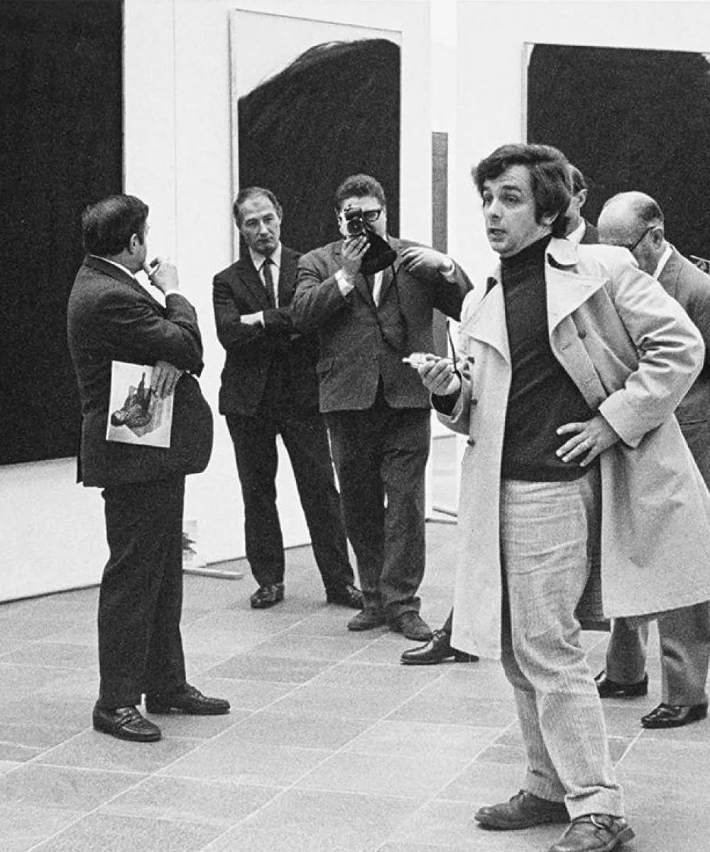 Group of men in suits at an art gallery, one man holds a camera, another gestures while speaking.