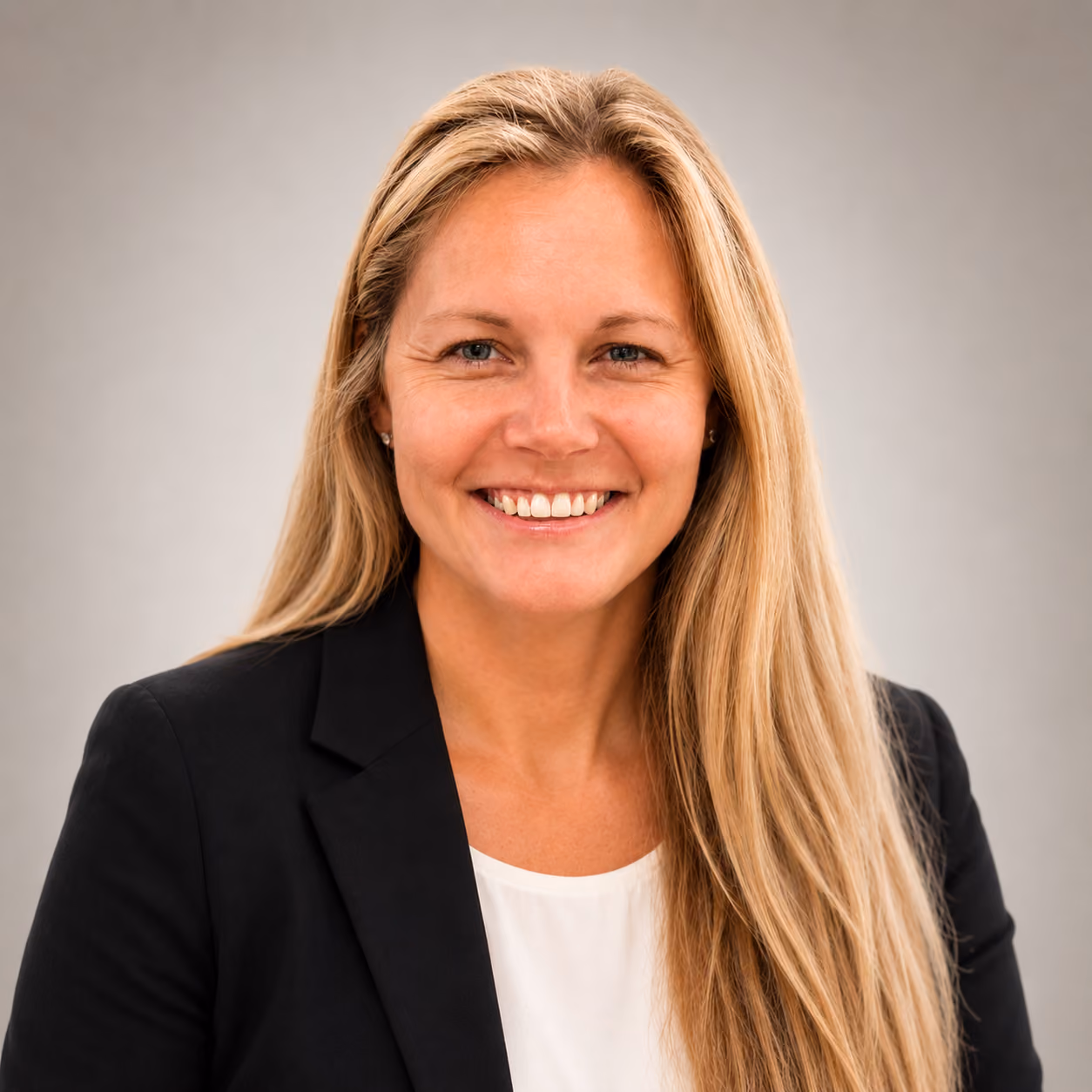 Smiling woman with long blonde hair wearing a black blazer and white top on a gray background.