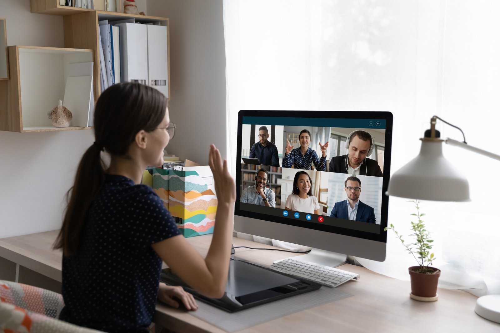 A woman waving to colleagues on a video call at her desk