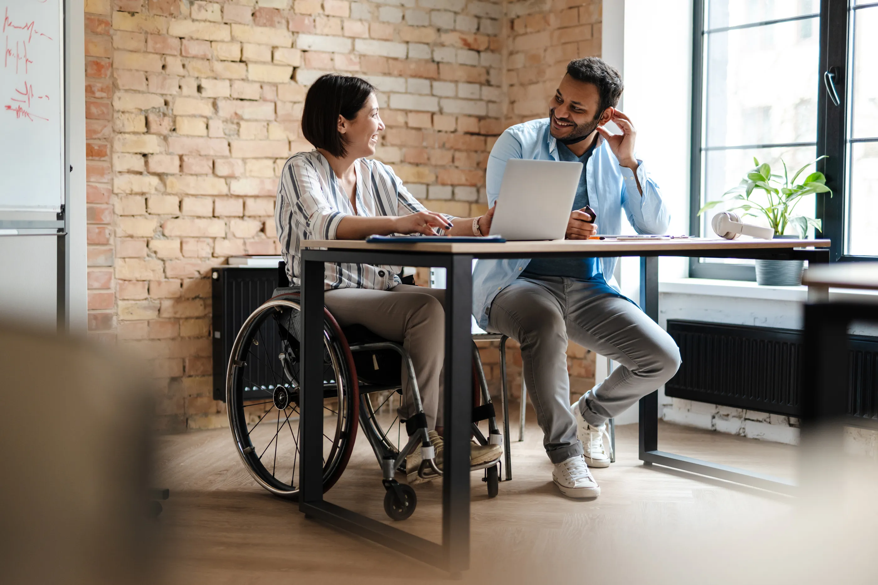 Two colleagues talking at a desk in a brick-walled office, one of them a wheelchair user
