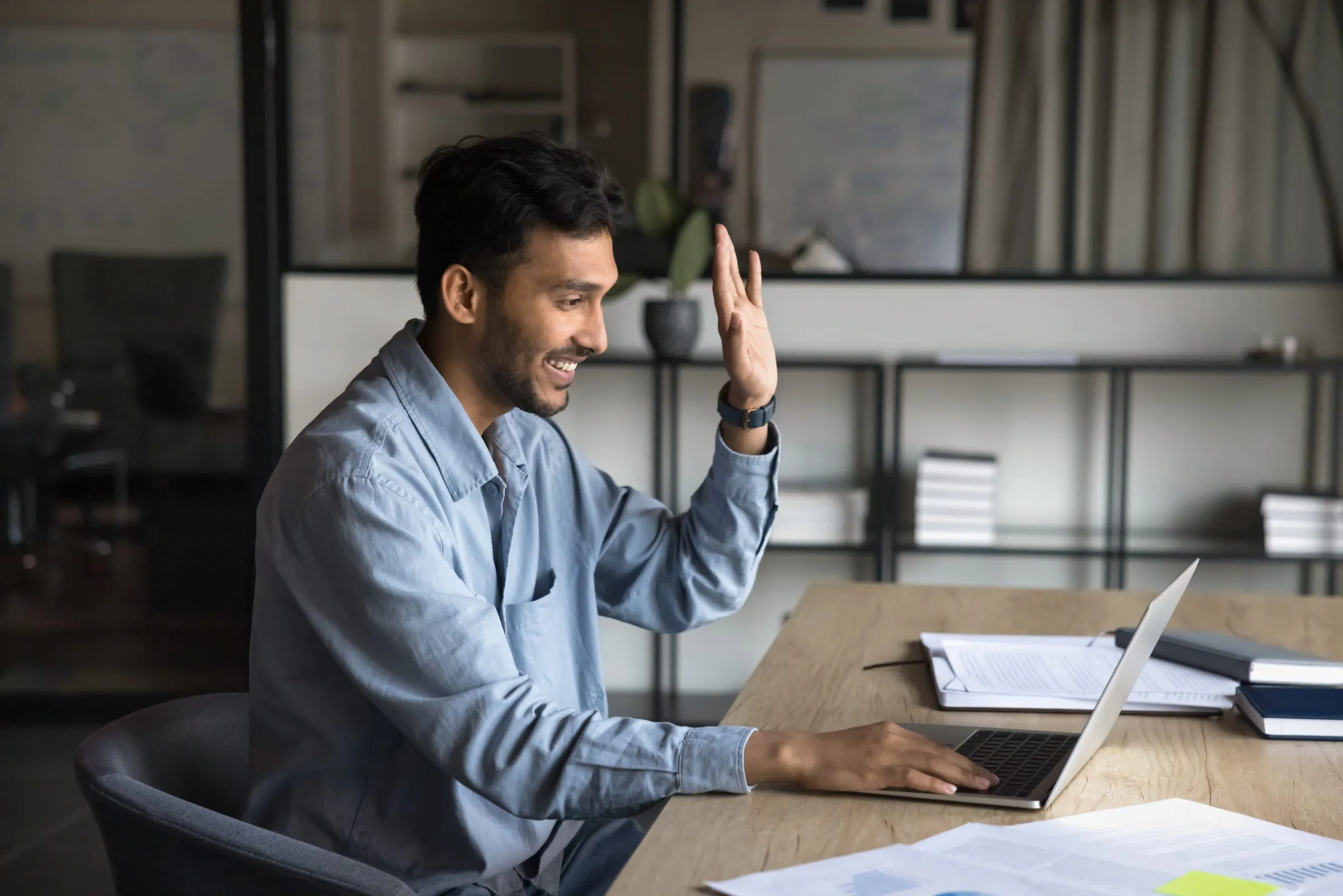 A man talking and gesturing at his desk, laptop open, in an open-plan office