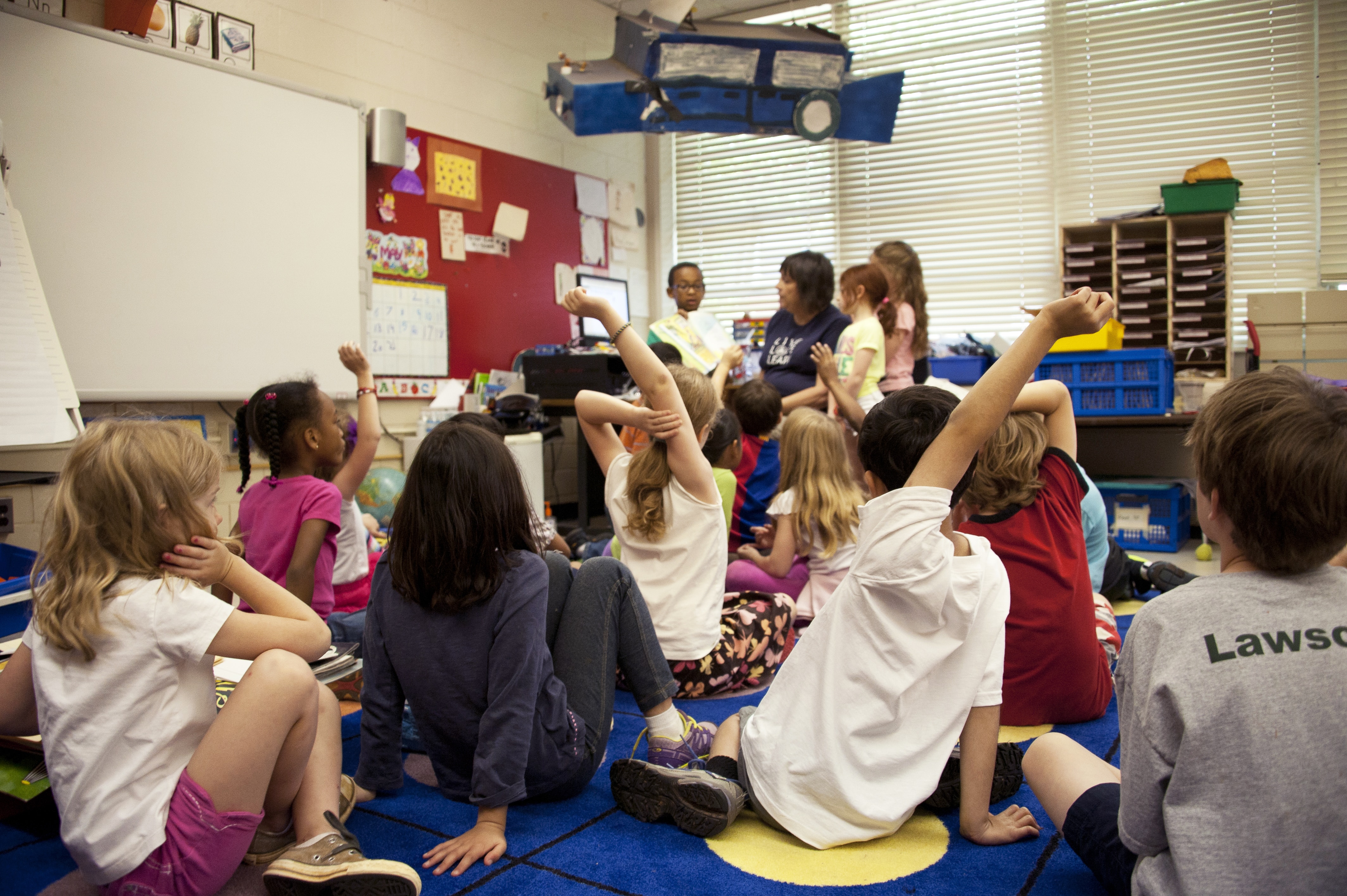 Elementary school students sitting on a colorful classroom rug, raising their hands while listening to a teacher read aloud from a storybook.