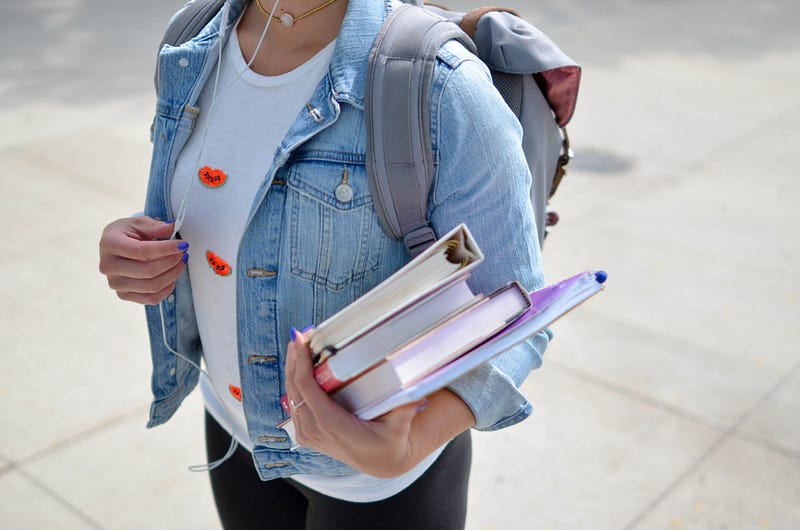 Mid-section view of a student carrying a stack of textbooks and notebooks, wearing a light-wash denim jacket, backpack, and wired earphones.