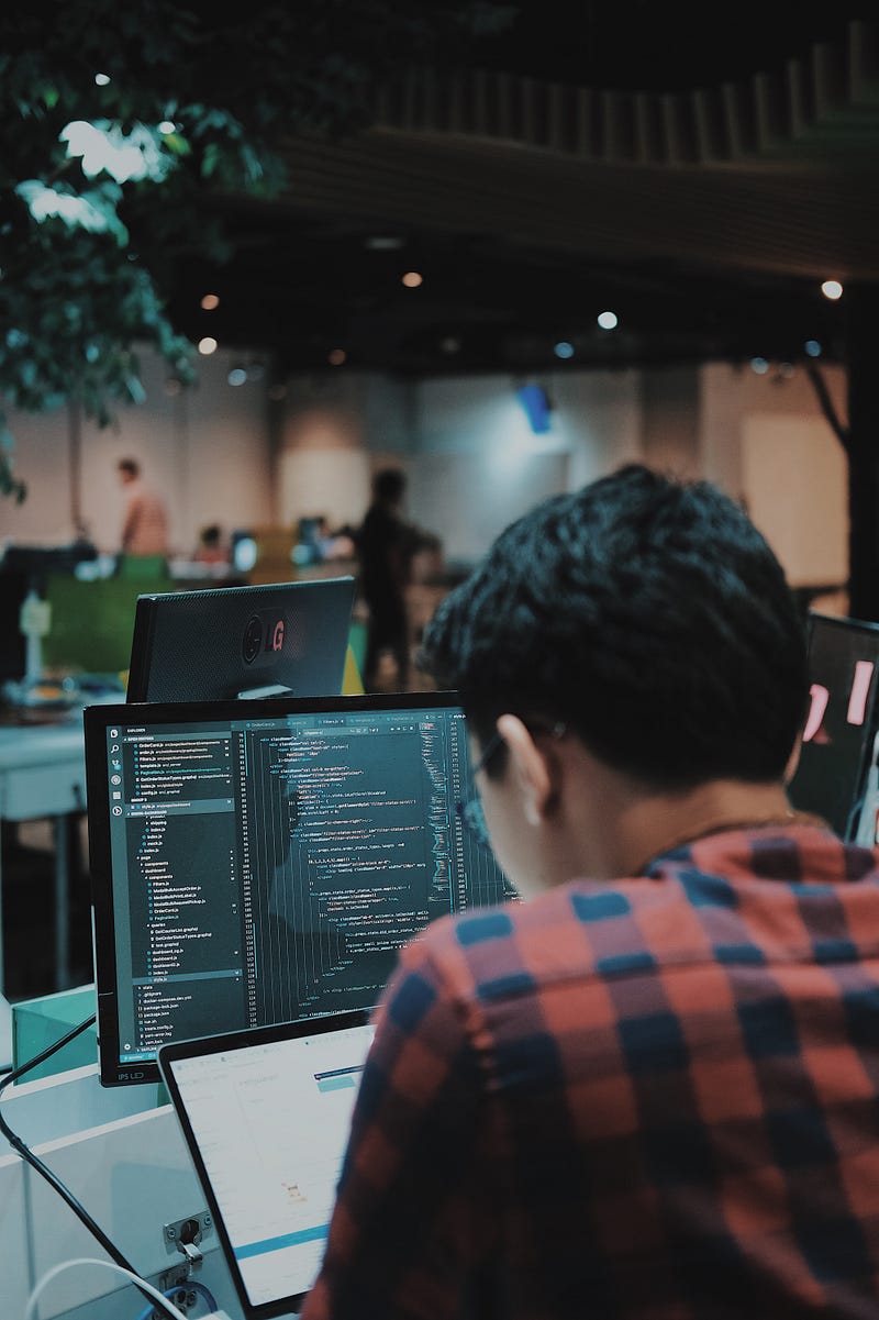 Over-the-shoulder view of a programmer in a red and black plaid shirt typing code on a desktop monitor and laptop in a modern office workspace.