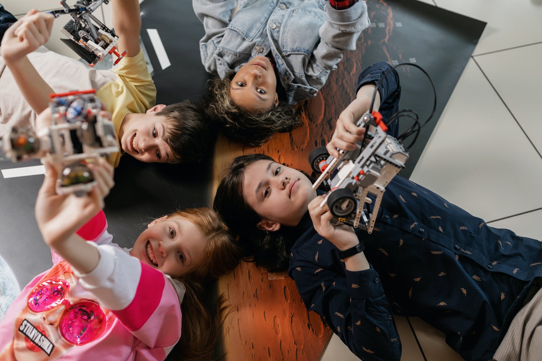 Top-down photograph of four young students lying on the floor in a head-to-head circle, smiling as they hold up built Lego robotics models toward the camera over a space-themed mat.