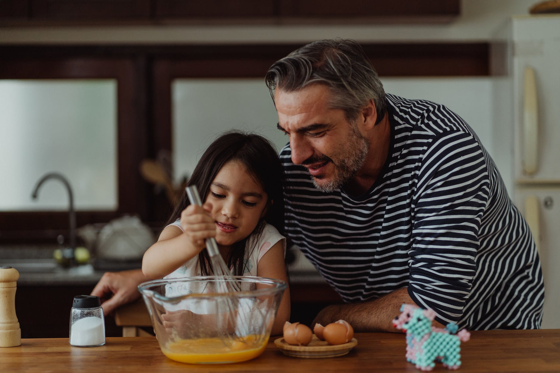 Father in a striped long-sleeve shirt smiling as he leans over to watch his young daughter carefully whisking eggs in a glass bowl on a kitchen counter.
