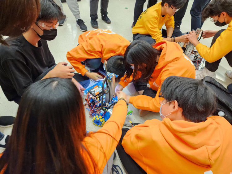 Group portrait of a robotics team wearing matching bright orange hoodies, safety glasses, and face masks posing together in a school gymnasium at a competition event.