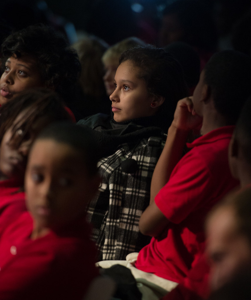 A young girl with her hair pulled back sits in an audience, looking attentively towards the front.