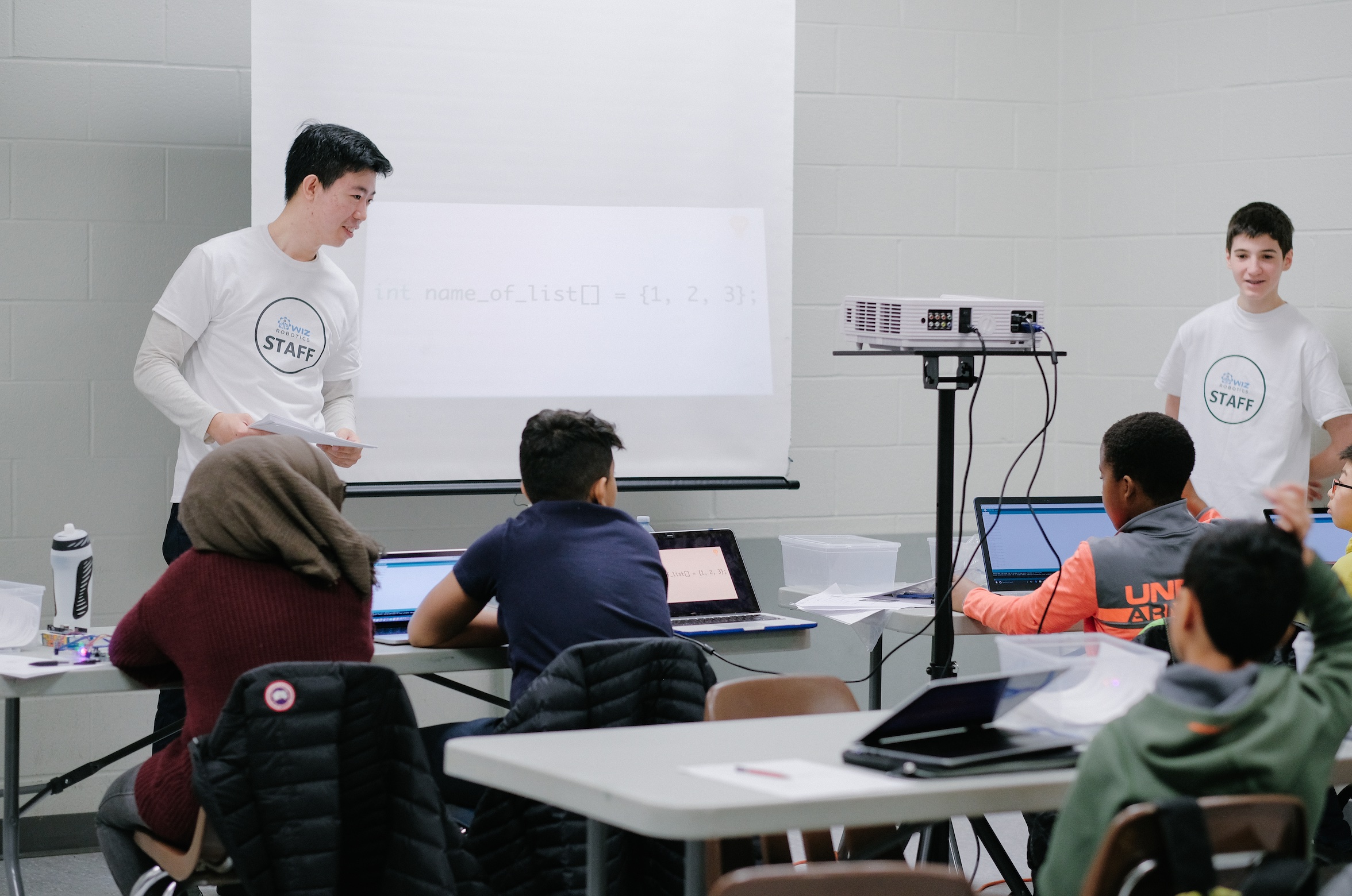 A Wiz Robotics staff member stands at the front of a classroom, teaching a group of students using a projector showing code.